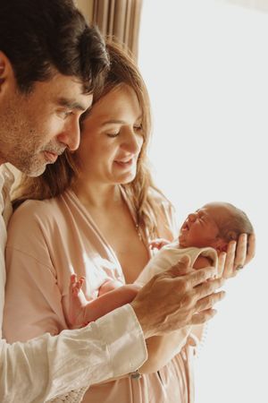 New parents lovingly cradling their sleeping newborn baby together near a bright window.