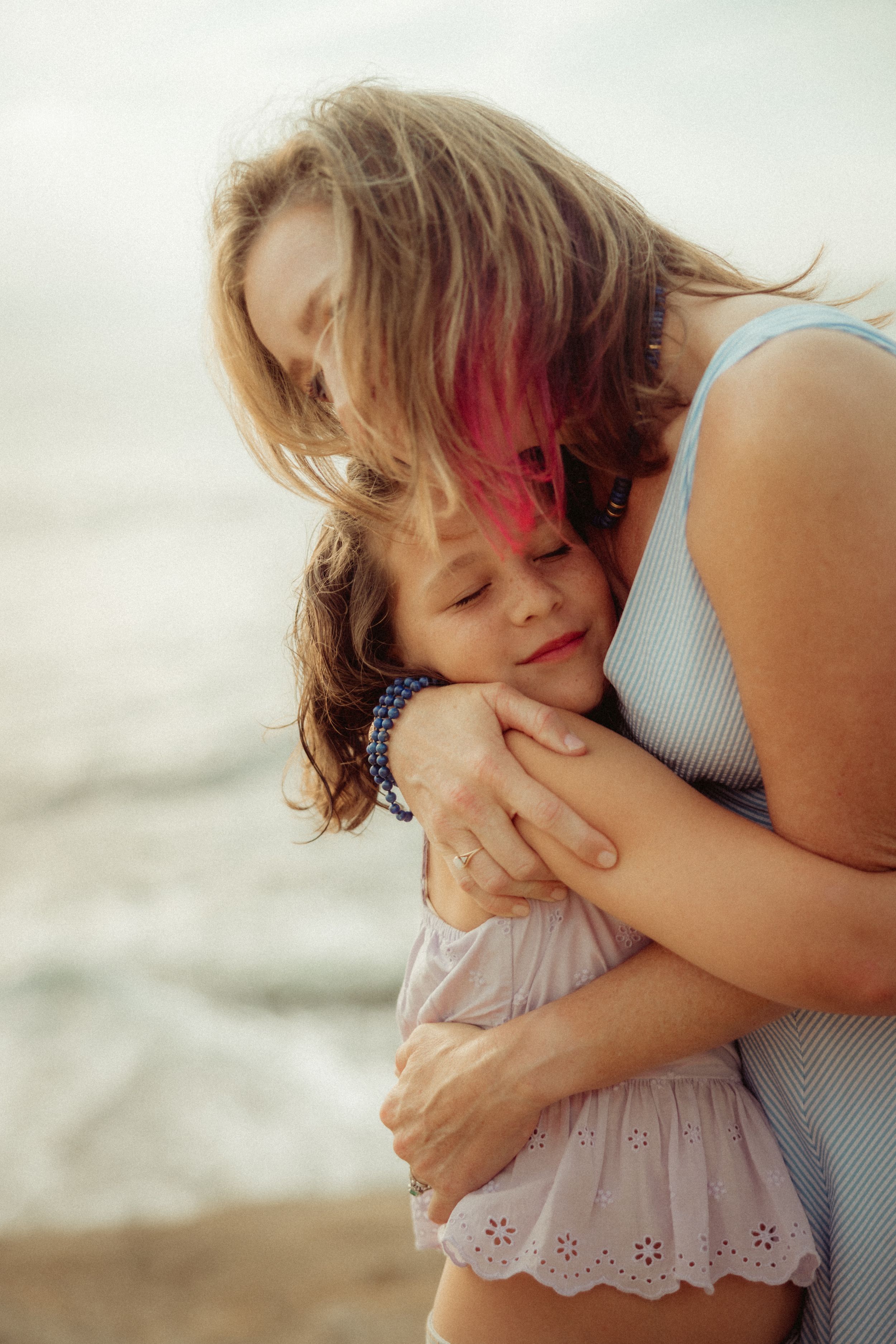 Mother with pink-highlighted hair tenderly embraces young daughter on a beach, both sharing a peaceful, loving moment.