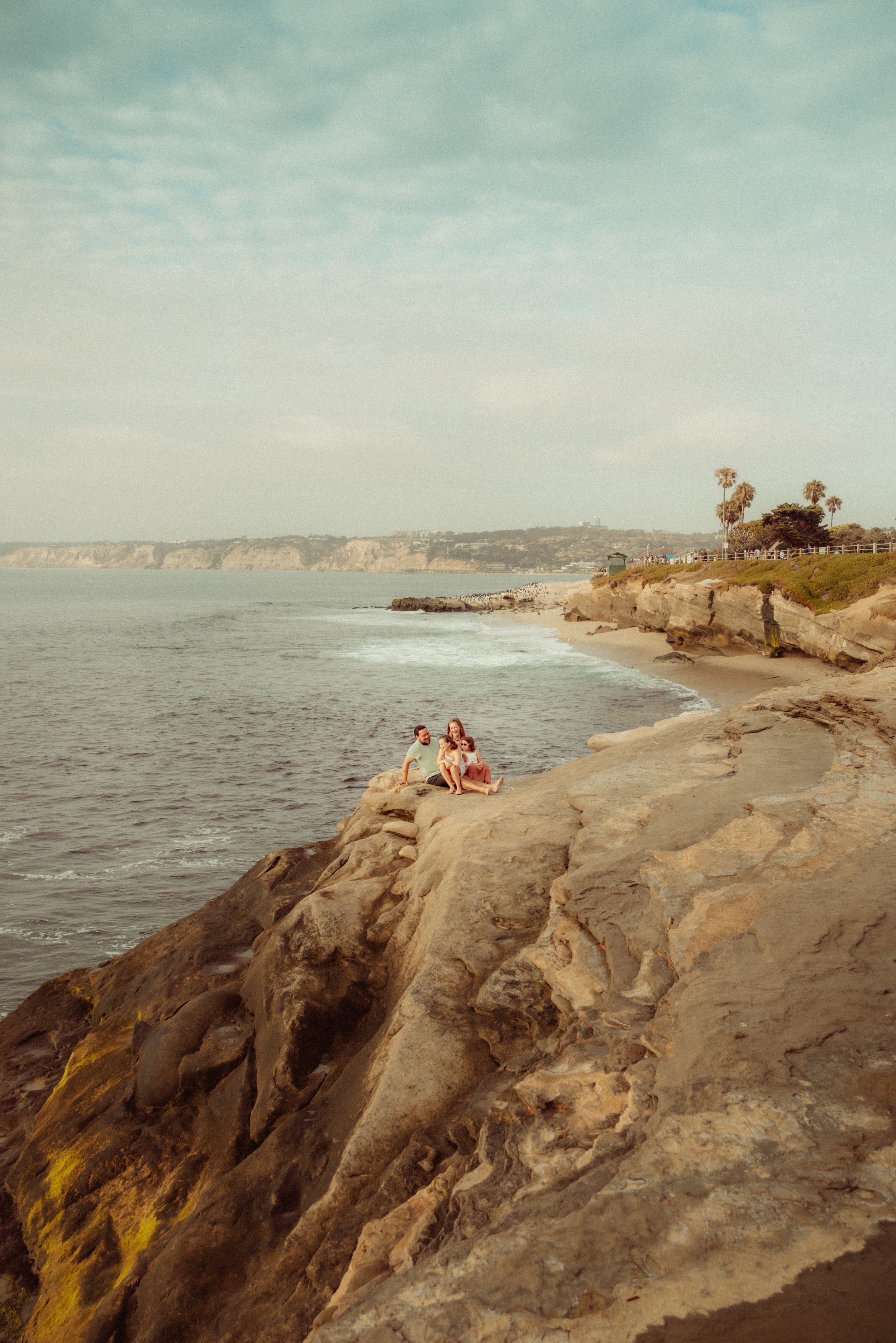 Two people sitting on rocky coastal cliffs overlooking the ocean with palm trees and blue skies in the background.