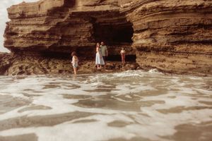 Family of three standing on a rocky beach cove with ocean waves and dramatic sandstone cliffs.