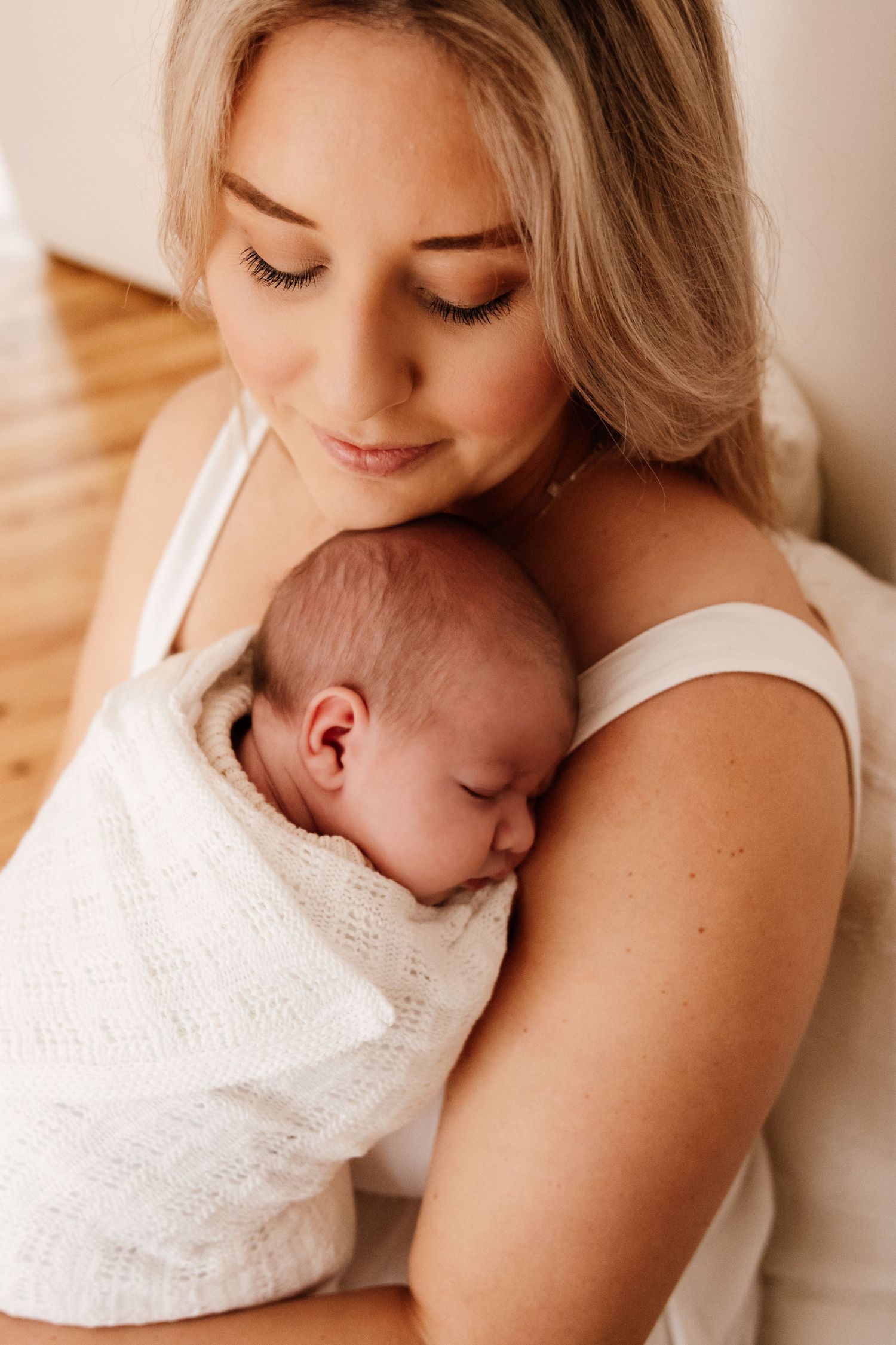Tender moment between parent and baby in white eyelet dress against neutral background.