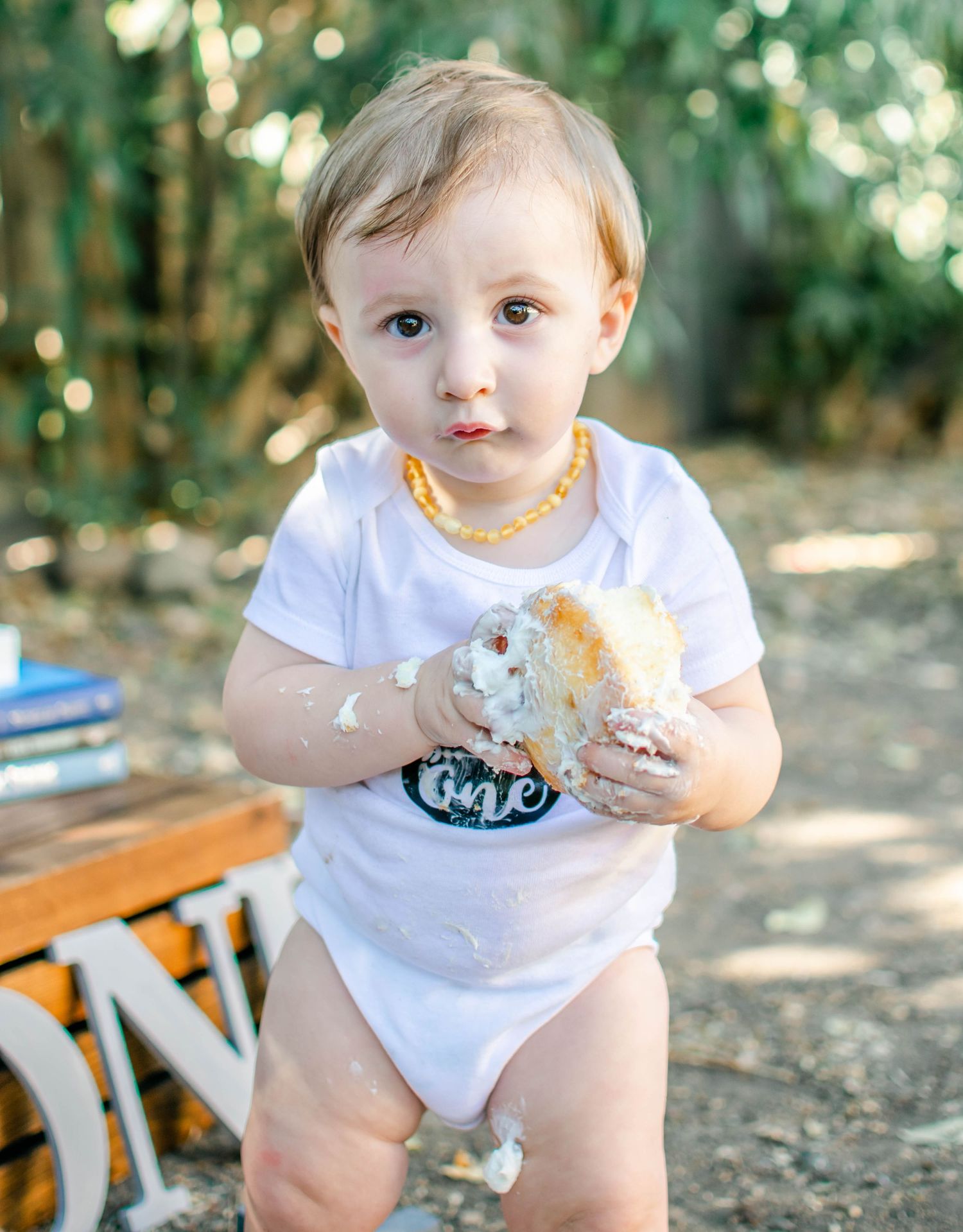 Baby in white onesie playing with cake during outdoor celebration photoshoot.