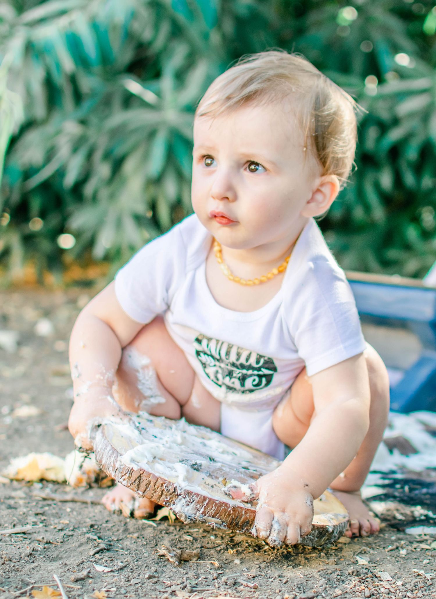 A small baby in a white t-shirt sits on sandy ground exploring and playing with the dirt.