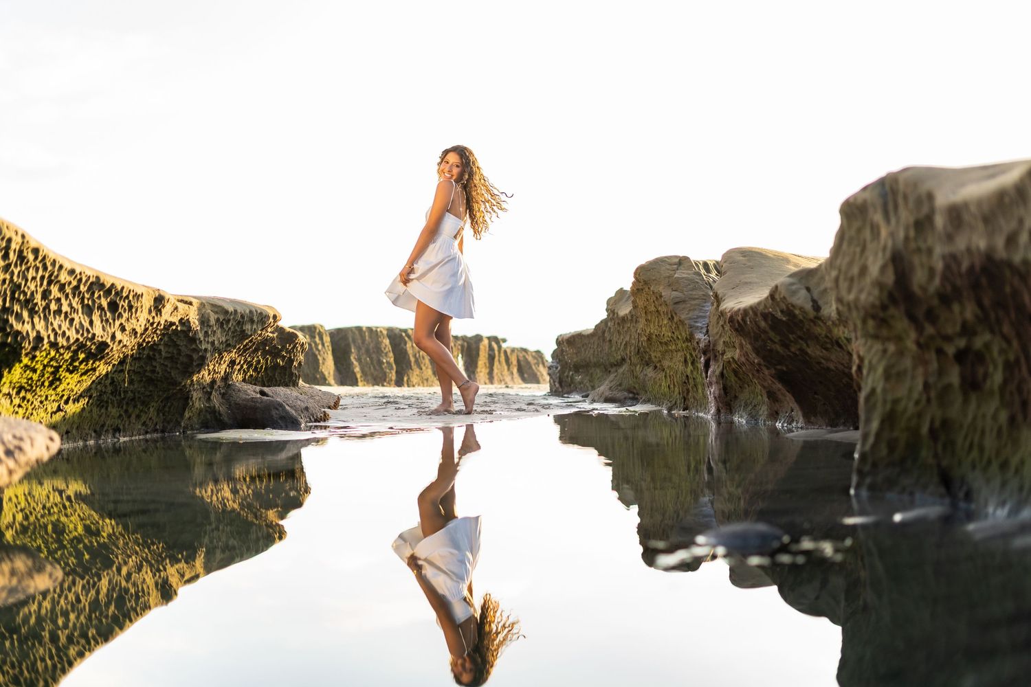 A high school senior in white dress leaps over reflective tide pool surrounded by coastal rock formations at sunset.
