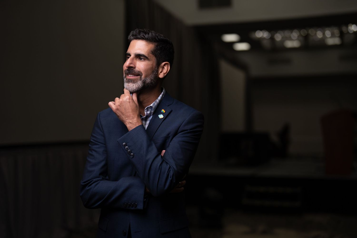 Confident man in navy suit poses thoughtfully with arms crossed in conference venue.