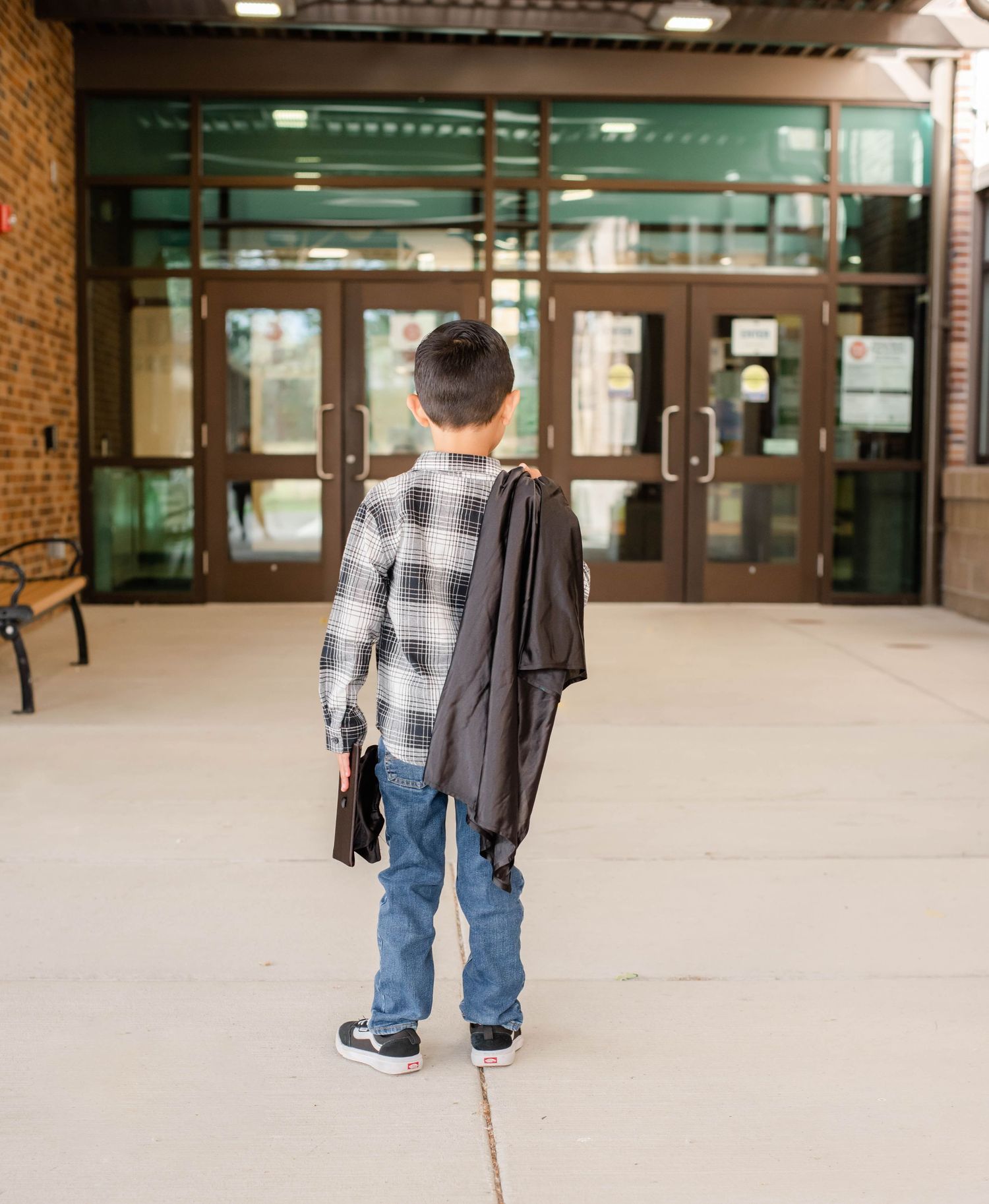 Graduate in plaid shirt and jeans stands in front of brick building entrance.