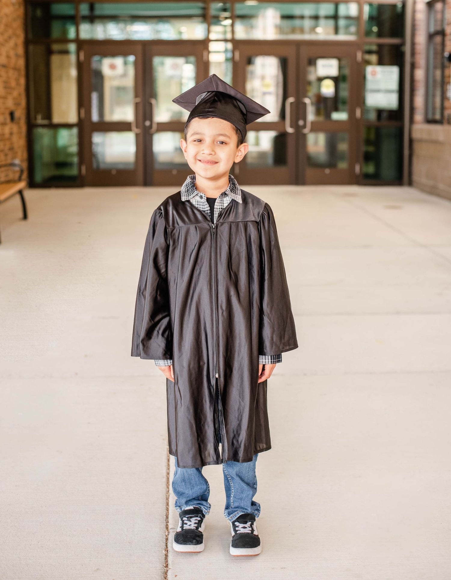 Graduate in black cap and gown poses in front of institutional building.