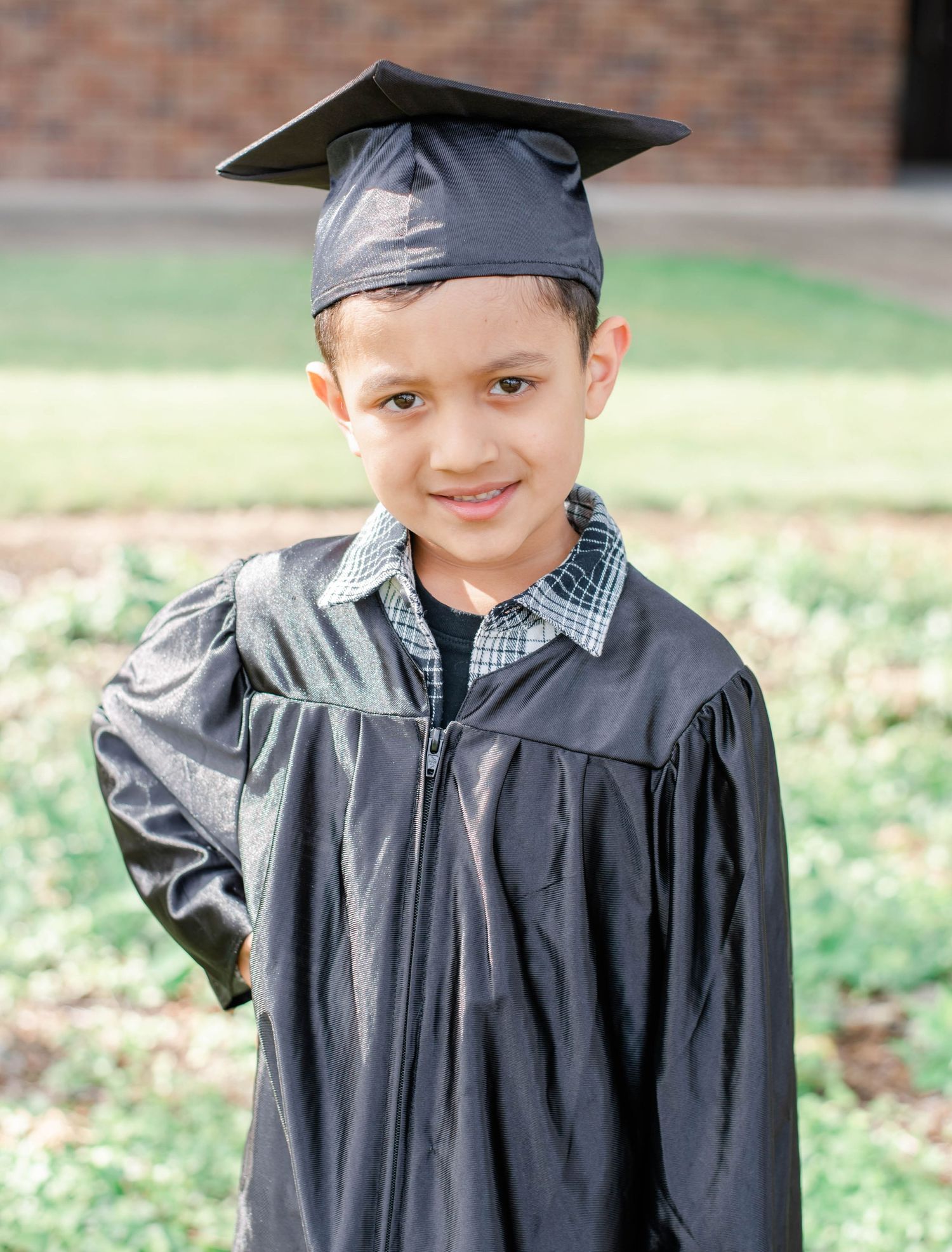 Young graduate in black cap and gown smiles outdoors near brick building.