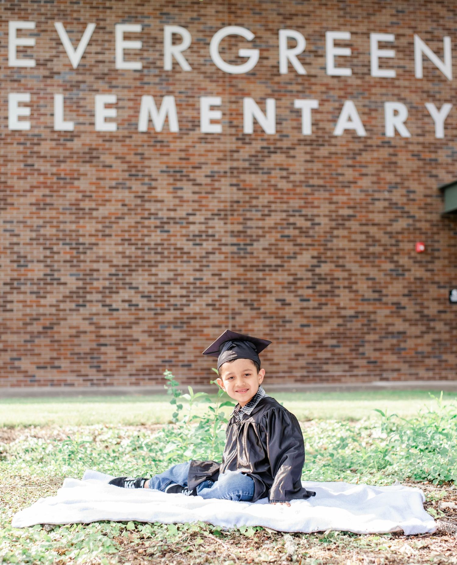 A graduate in a black cap and gown poses in front of Evergreen Elementary School's brick facade.