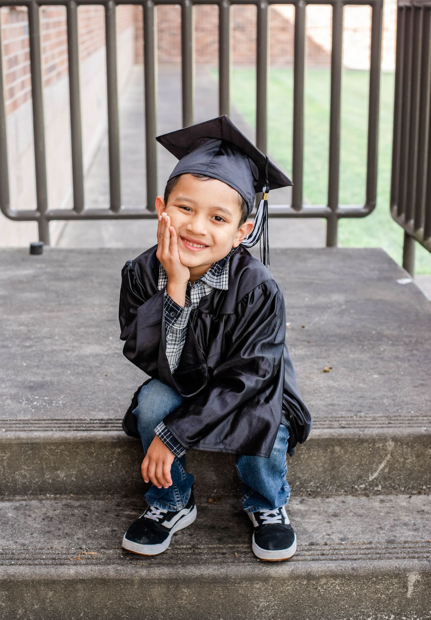 A graduate in black cap and gown crouches on concrete steps while posing for photos.