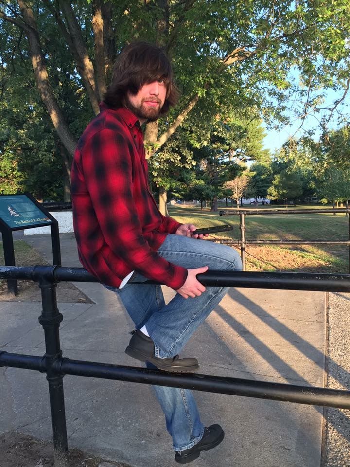 Person in red plaid flannel and jeans sits casually on metal railing in park setting.