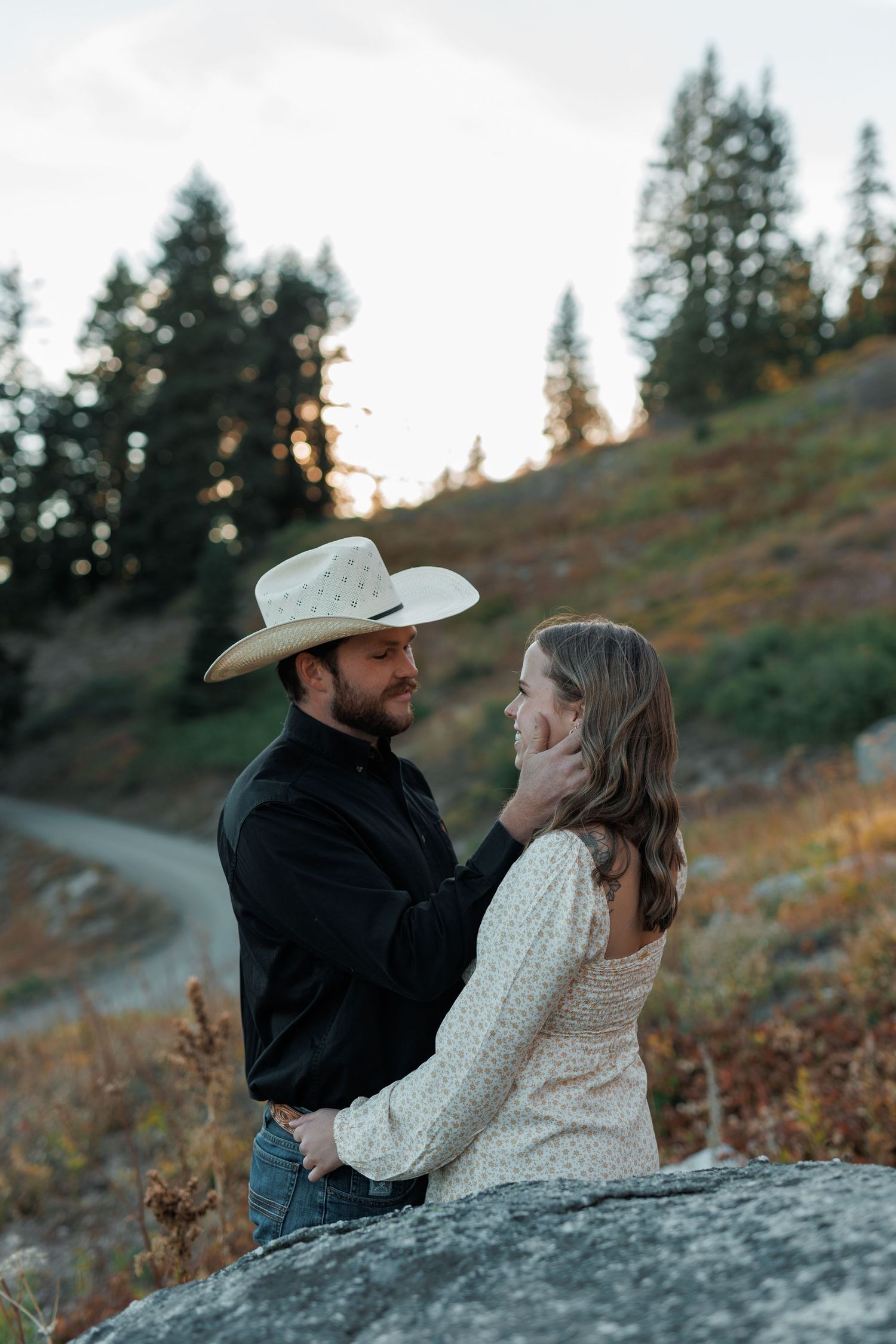 A couple shares an intimate moment standing by a rock at sunset with evergreen trees in the mountain background.