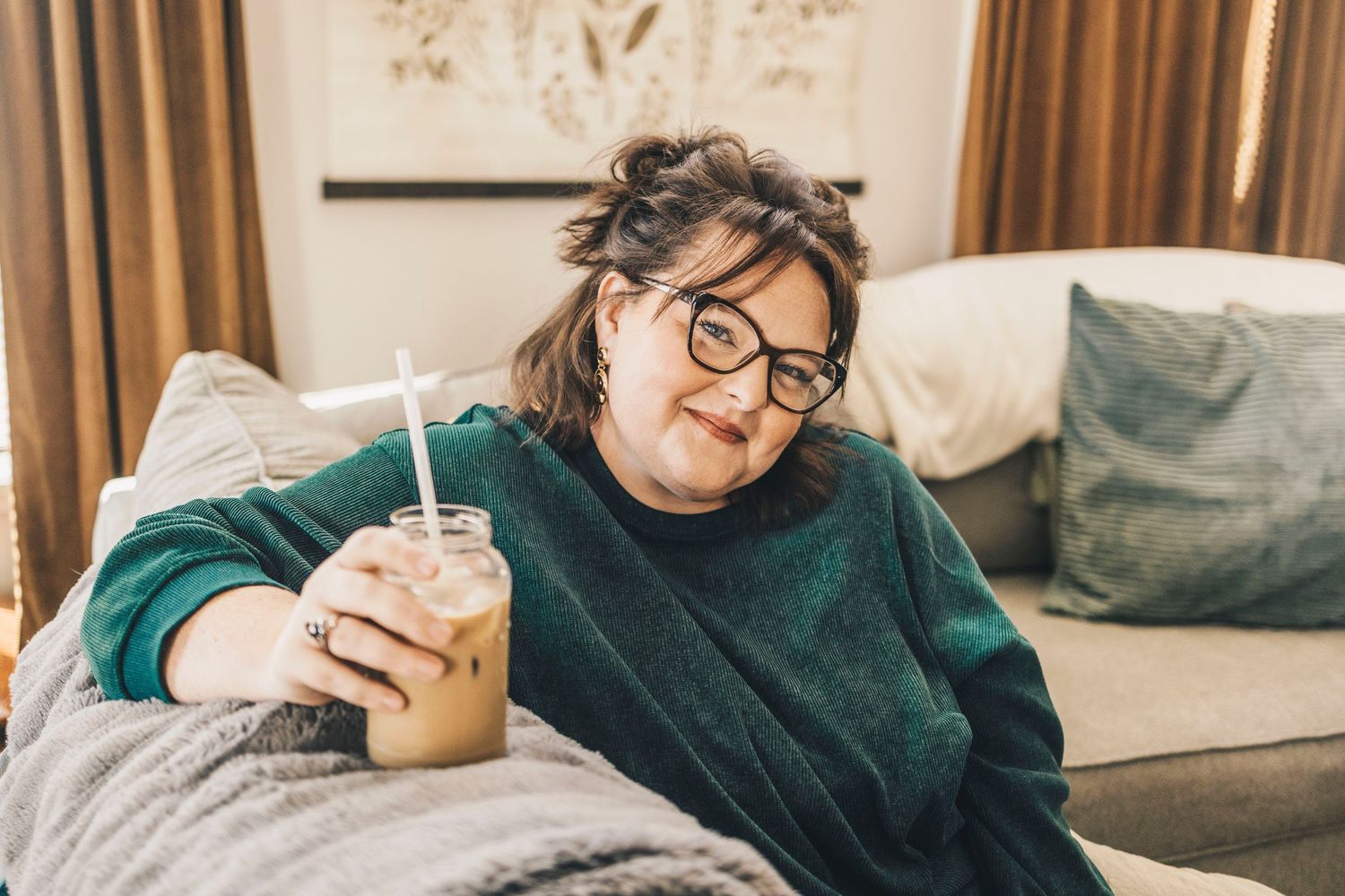 Woman in green sweater relaxing on couch holding iced coffee with straw.