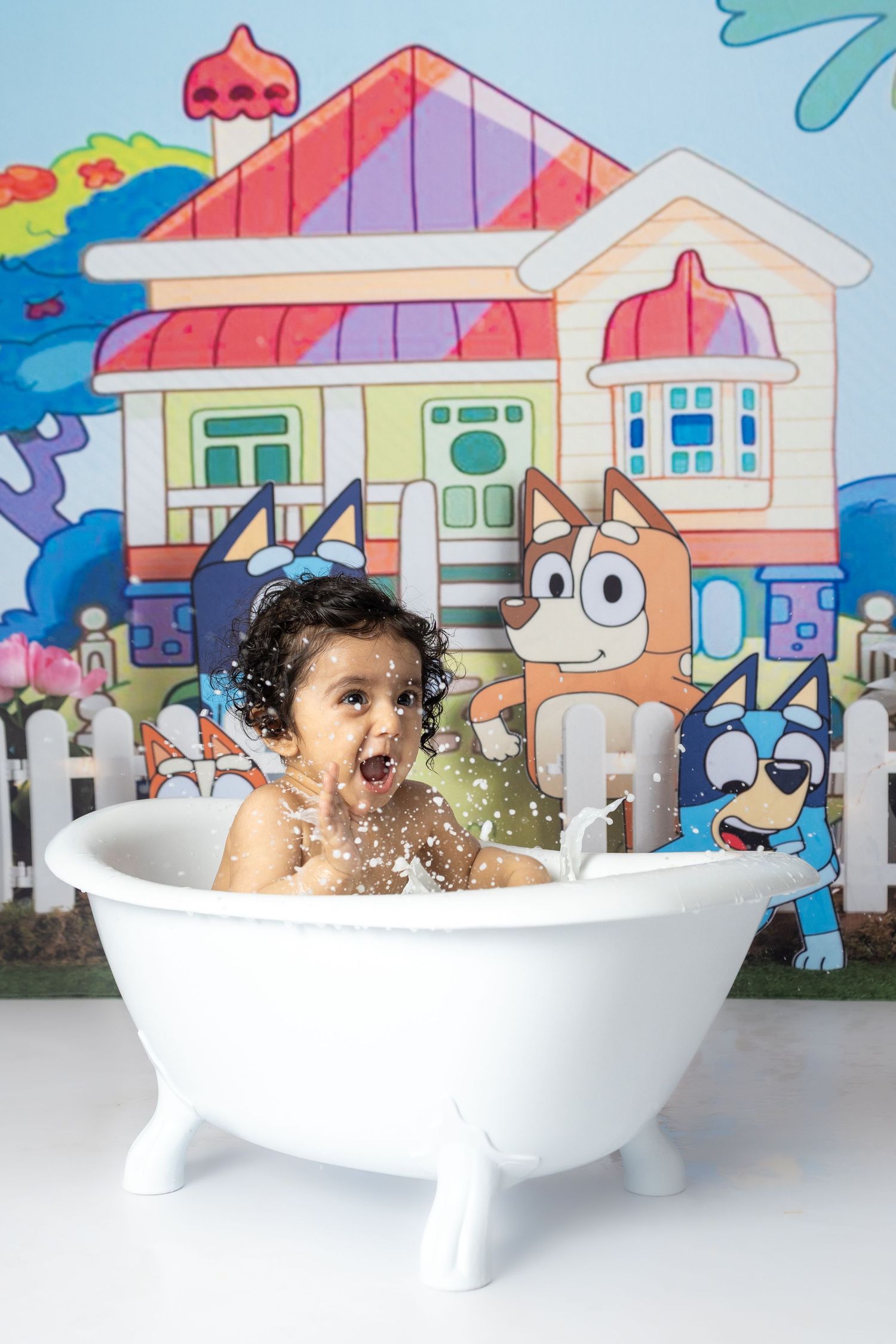 A playful cake smash photo session in a white bathtub with splashing water and colorful backdrop.