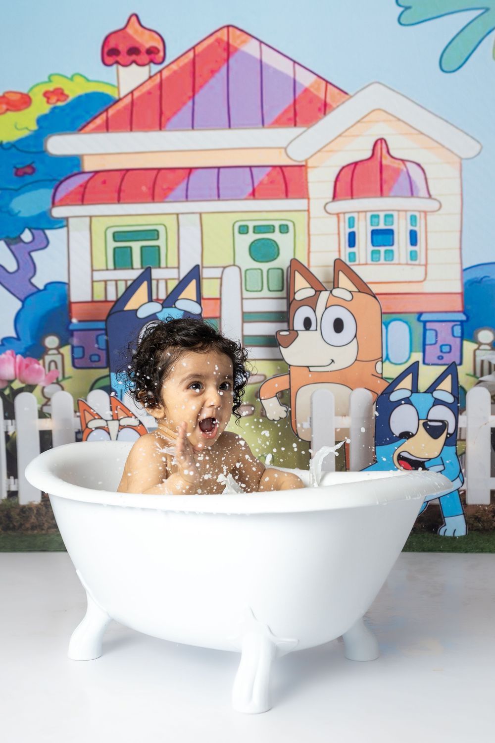 A playful cake smash photo session in a white bathtub with splashing water and colorful backdrop.