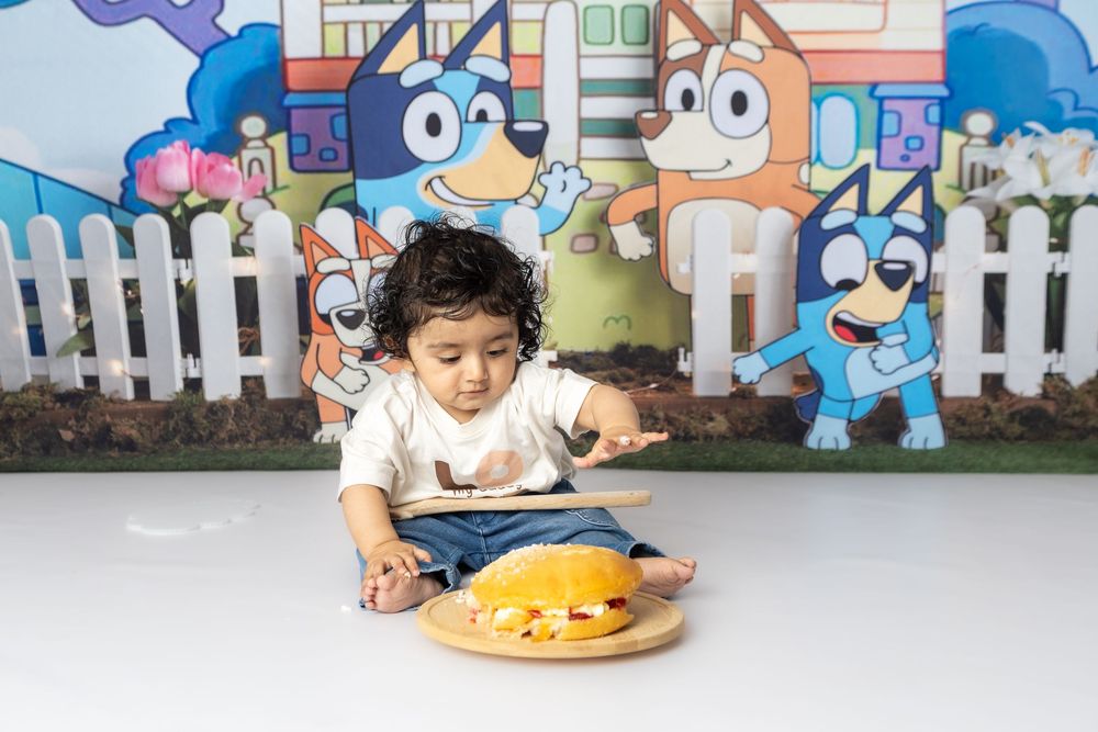 A child plays with a cake smash setup against a colorful cartoon background with white picket fence.