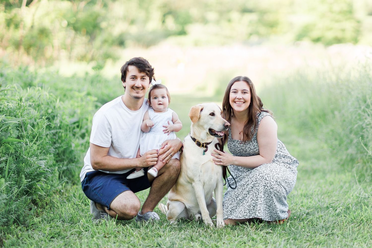 A couple and their baby pose outdoors with their friendly golden retriever on a sunny afternoon.