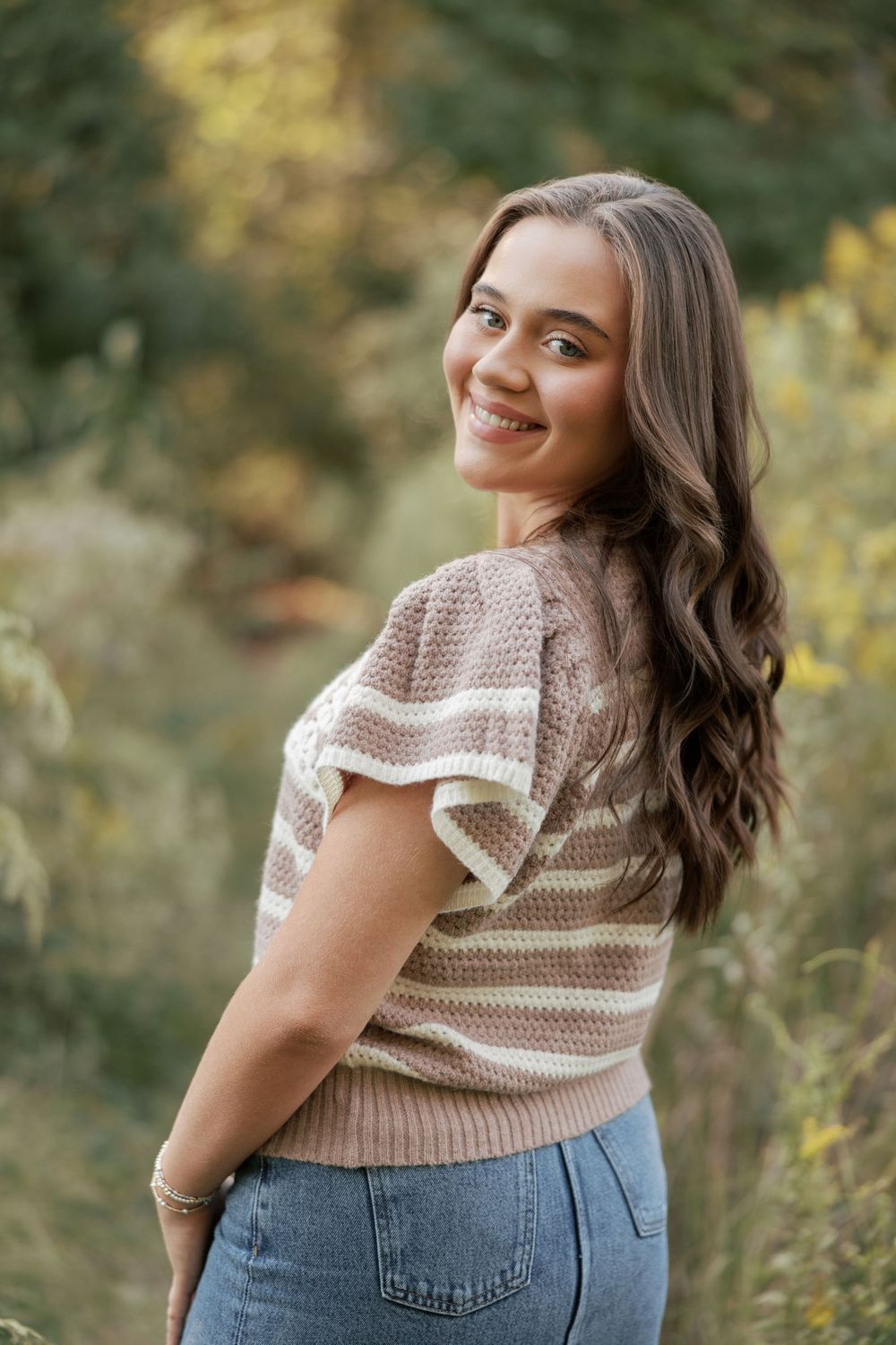 A person wearing a beige knit sweater with white trim and blue jeans poses outdoors against a blurred natural background.