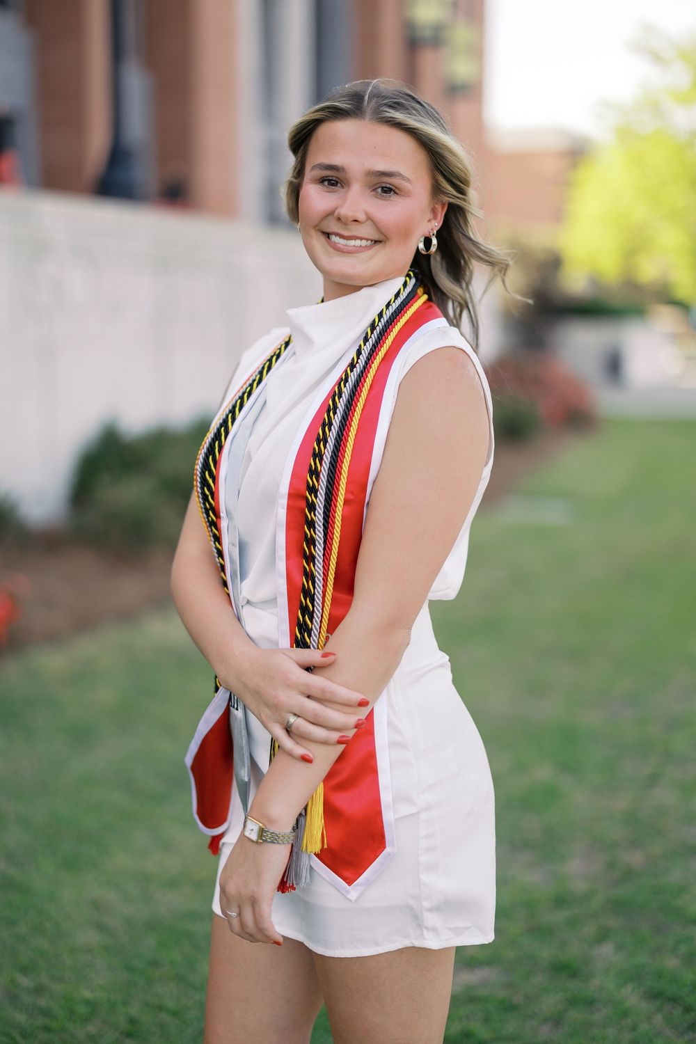 Graduate wearing white outfit with orange and black graduation stoles stands on grass near brick building.