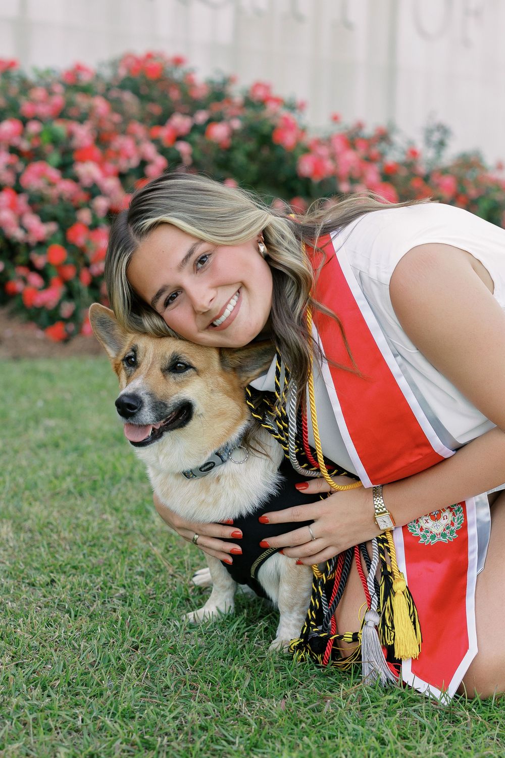 A cheerleader in red and white uniform poses playfully with a tan mixed-breed dog against a backdrop of pink flowers.