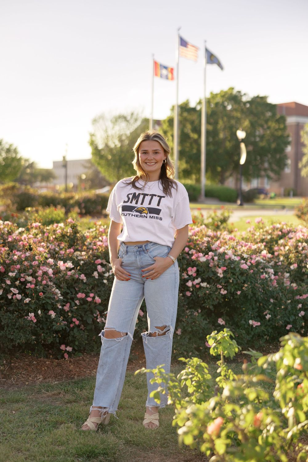A person wearing ripped jeans and a white t-shirt poses in front of a garden with blooming pink roses.