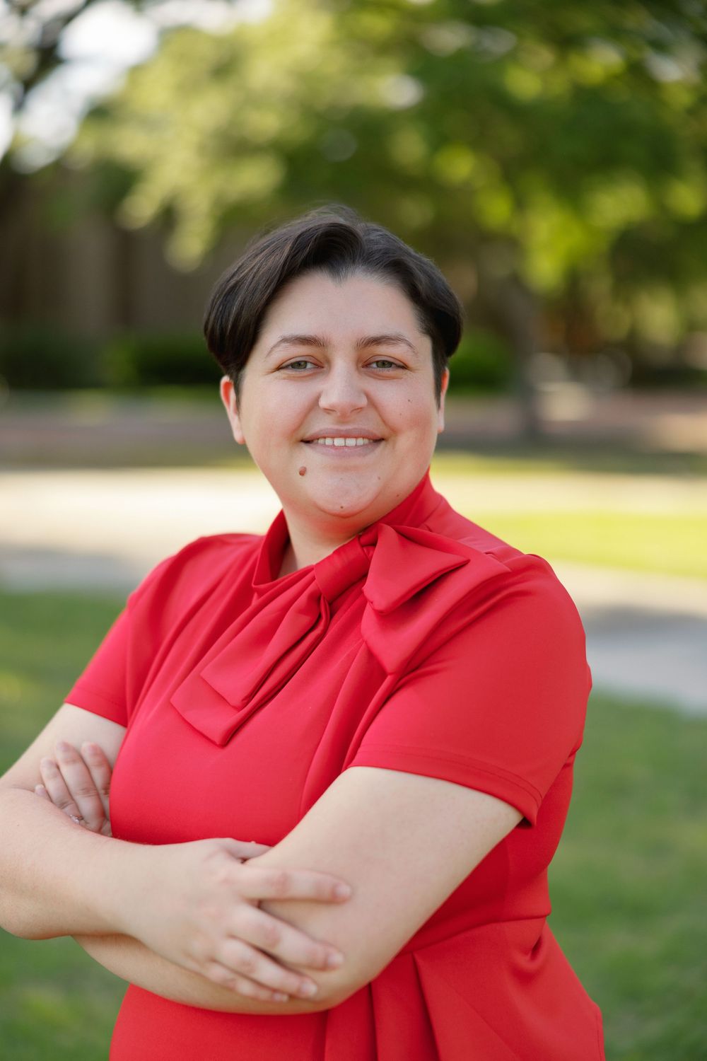 A person in a bright red shirt with a bow collar stands with crossed arms outdoors in a park-like setting.