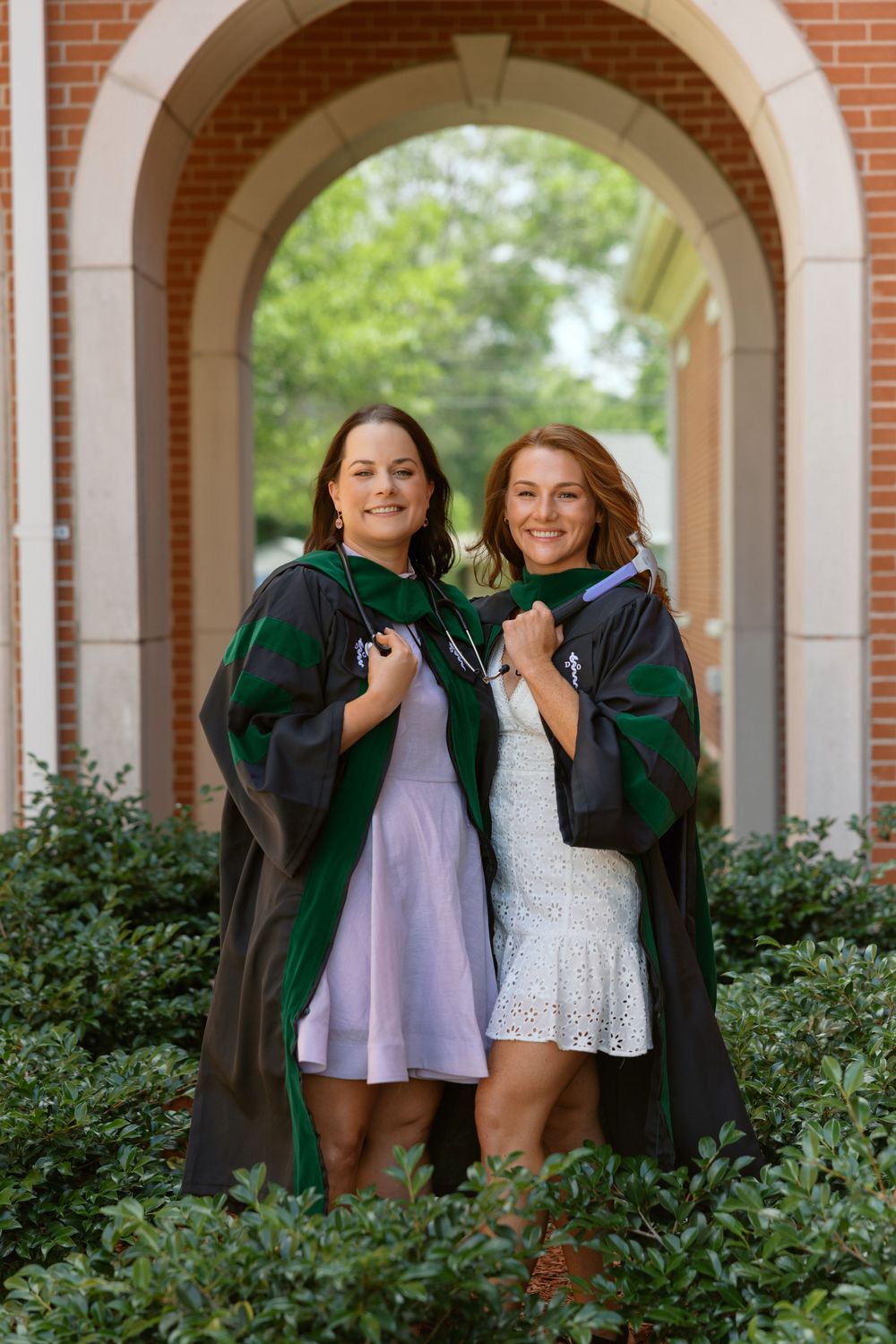 Two graduates in green caps and gowns share a joyful moment under brick archways on their graduation day.