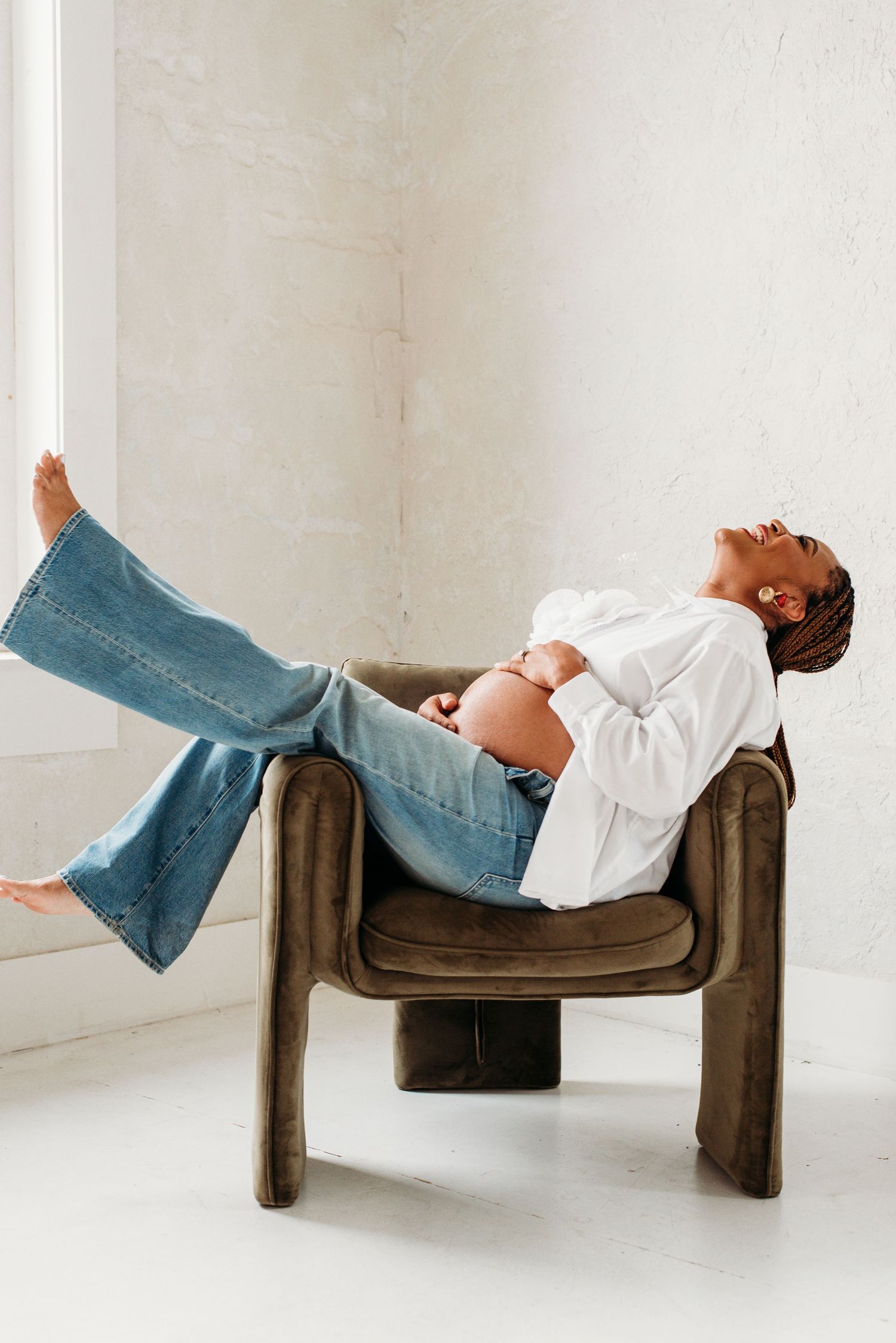 Smiling pregnant woman in ripped jeans sitting on kitchen counter with open belly showing.
