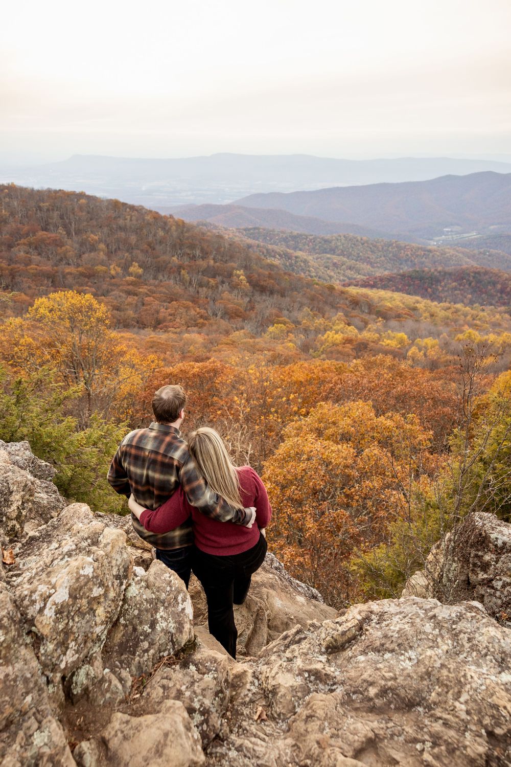 Two hikers enjoy an autumn mountain vista from a rocky overlook as fall colors paint the rolling hills in golden hues.