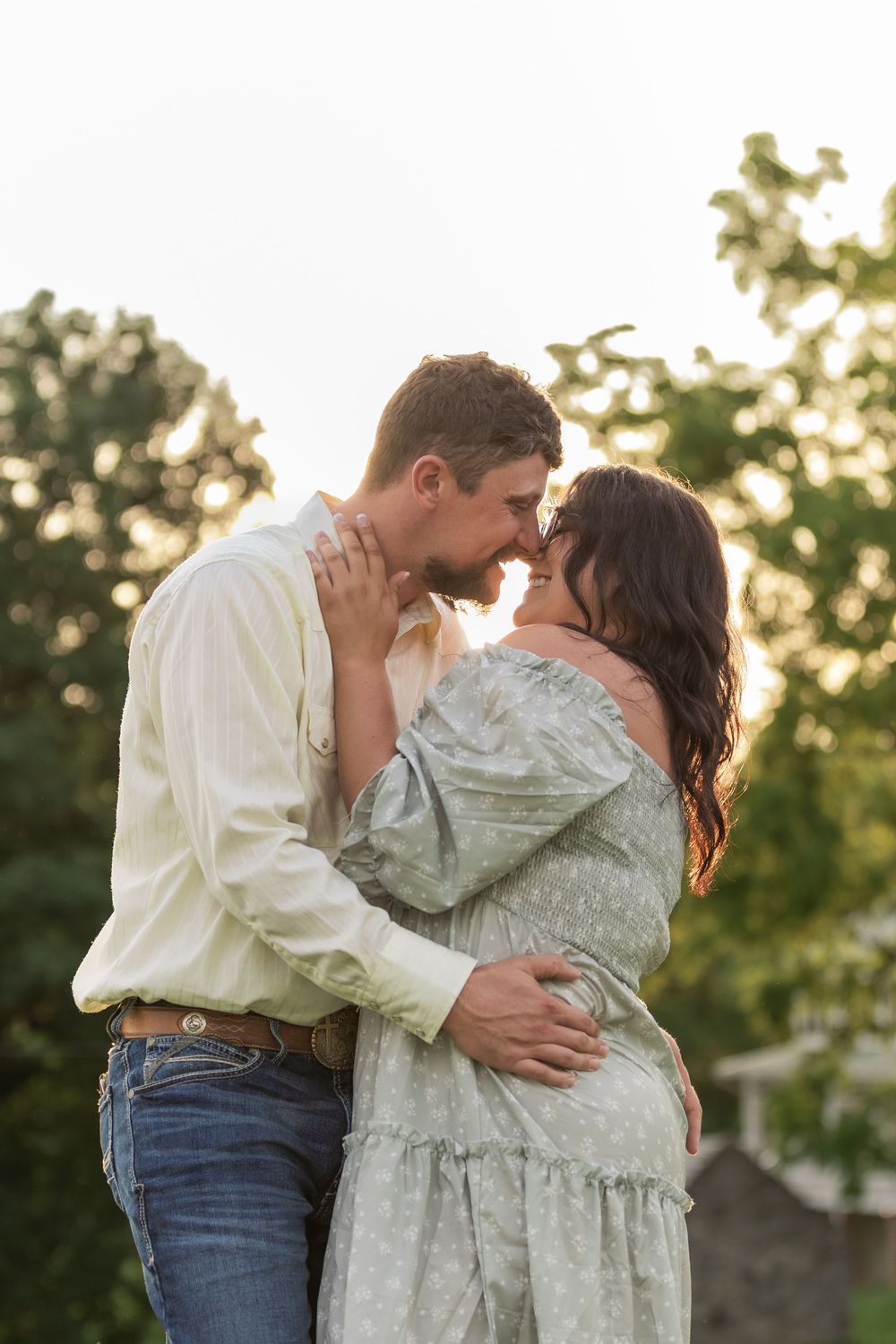 A romantic couple embraces at sunset in a rustic outdoor setting wearing matching cream and sage green outfits.
