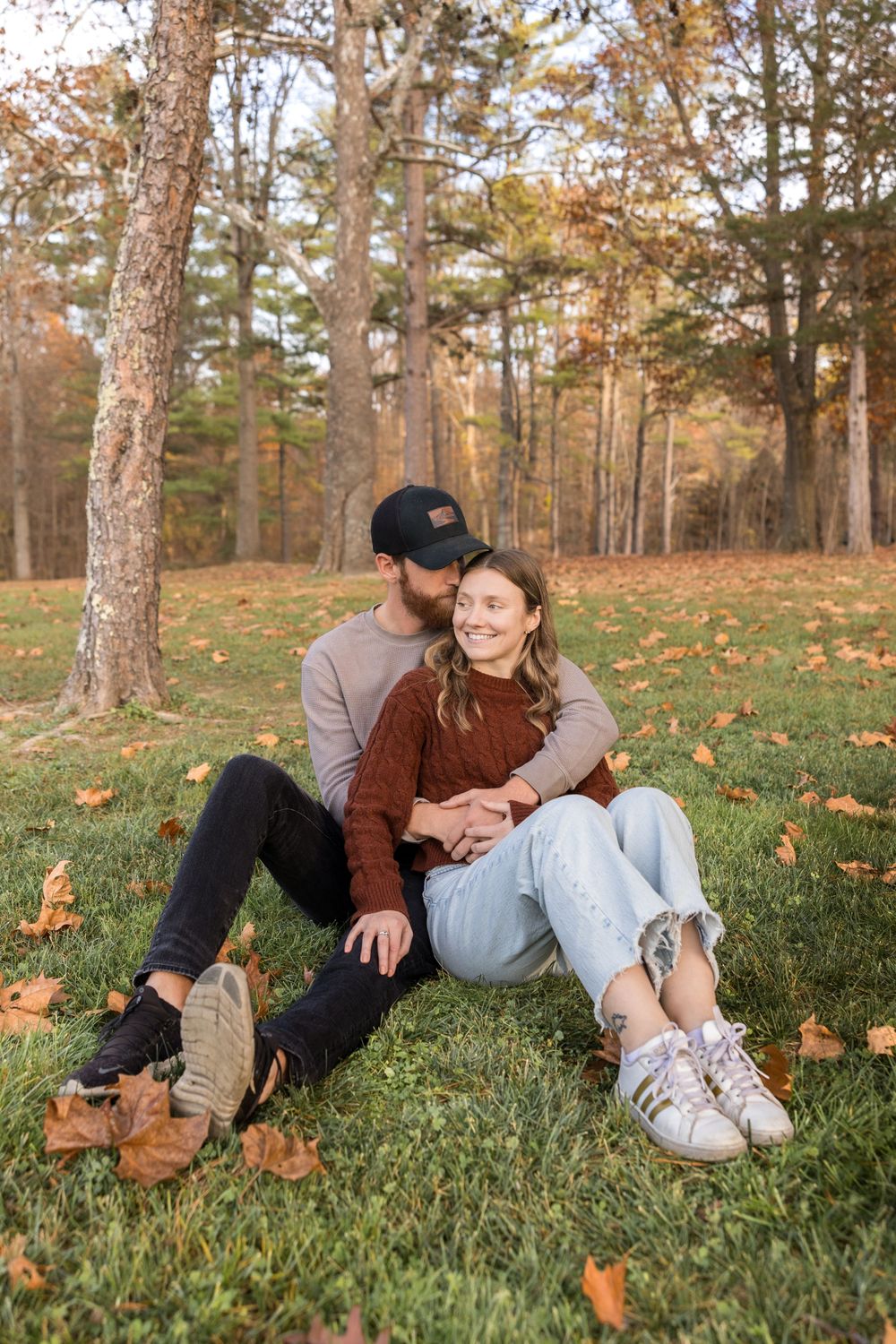 Two people sitting together on grass surrounded by fall foliage and trees in an autumn outdoor setting.