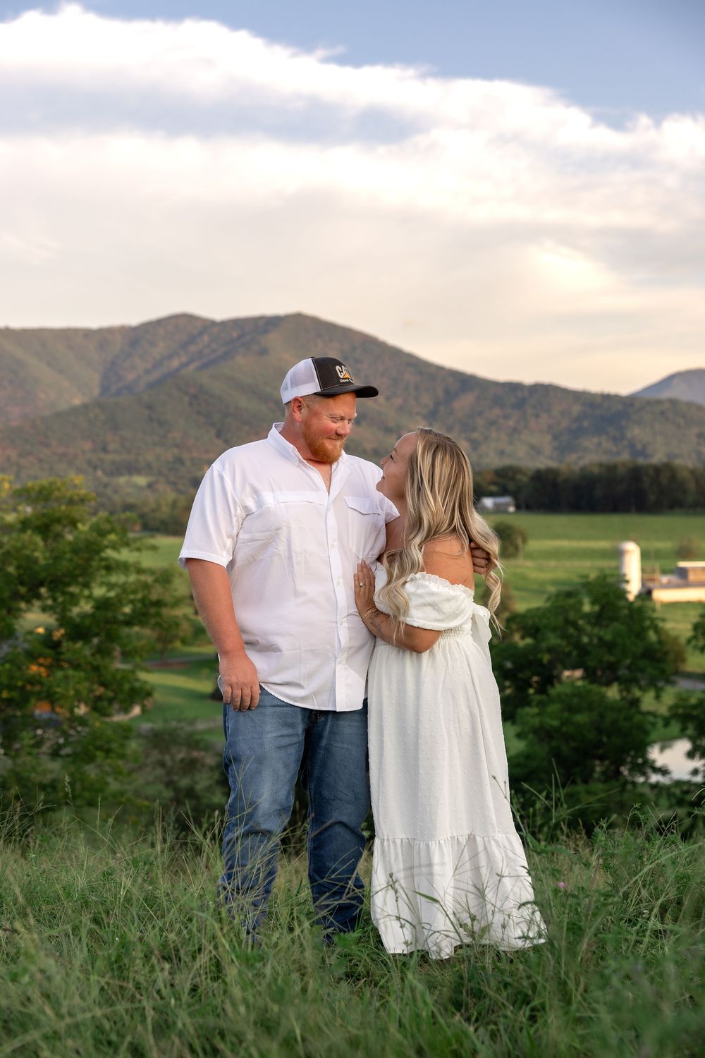 A couple stands together in a grassy field with mountains in the background at sunset.