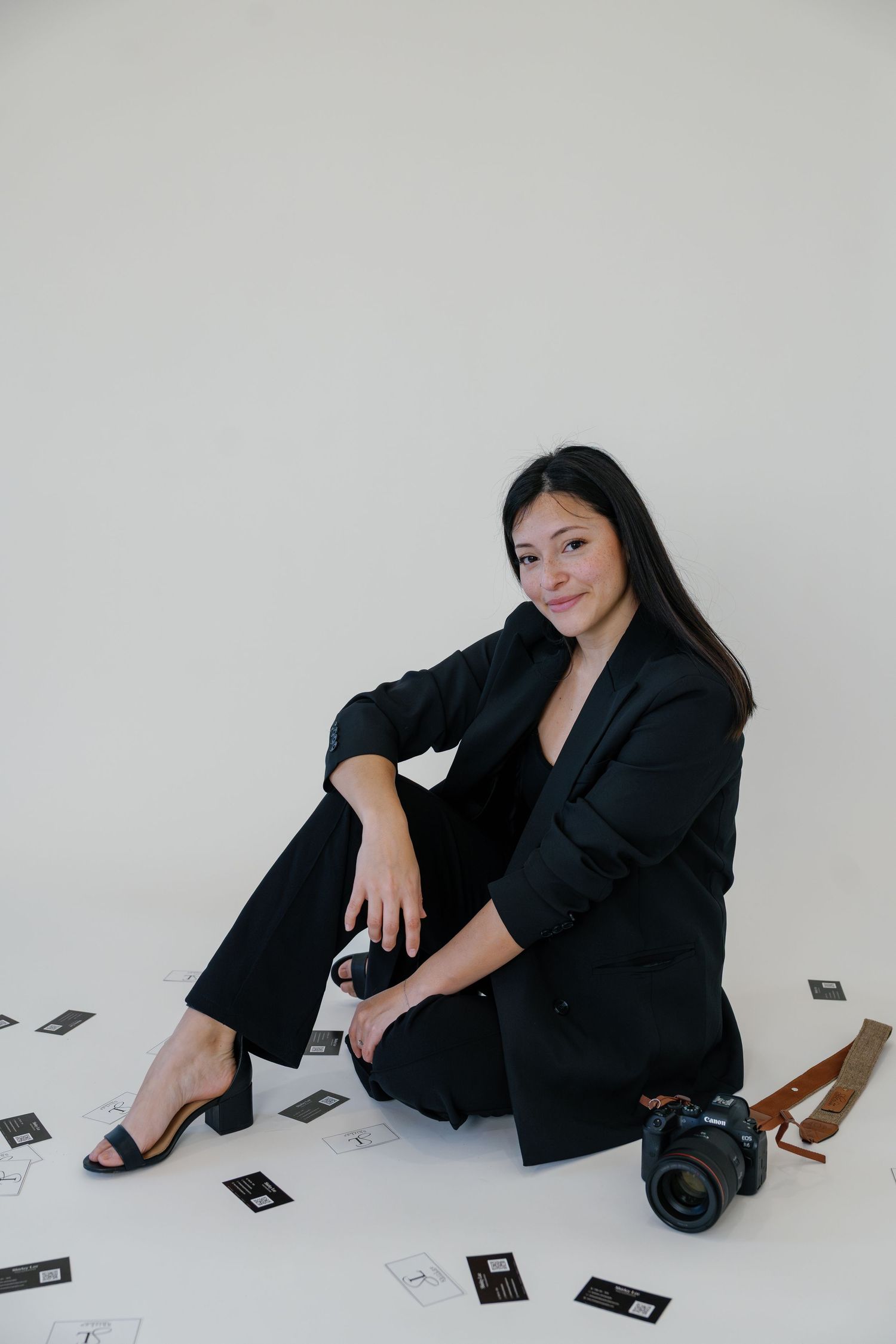 Professional wearing a black blazer and pants sits on floor with camera and scattered polaroid photos in minimalist setting.