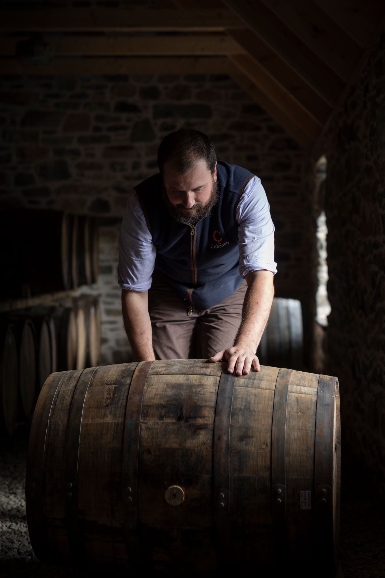 A worker in a vest inspects and rolls wooden whiskey barrels in a dimly lit distillery warehouse.