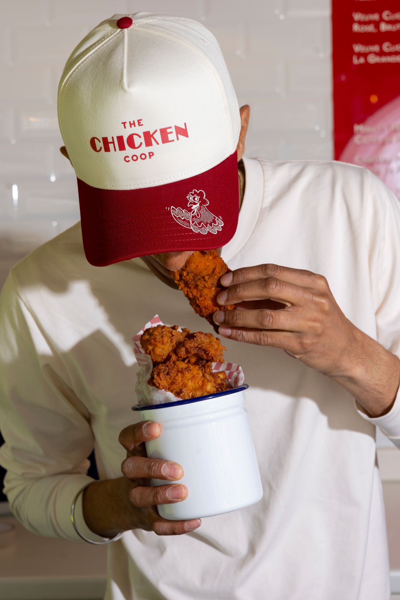 Someone wearing a red and white 'The Chicken Coop' baseball cap eating fried chicken from a white container.