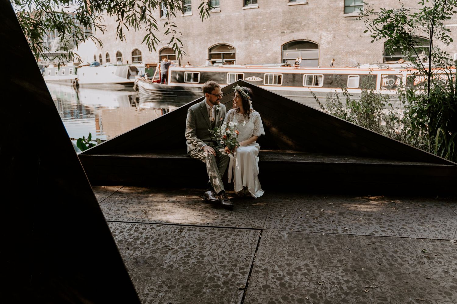 a couple are sat on a bench with a canal in the background. they are at camley nature reserve in London, kings cross.