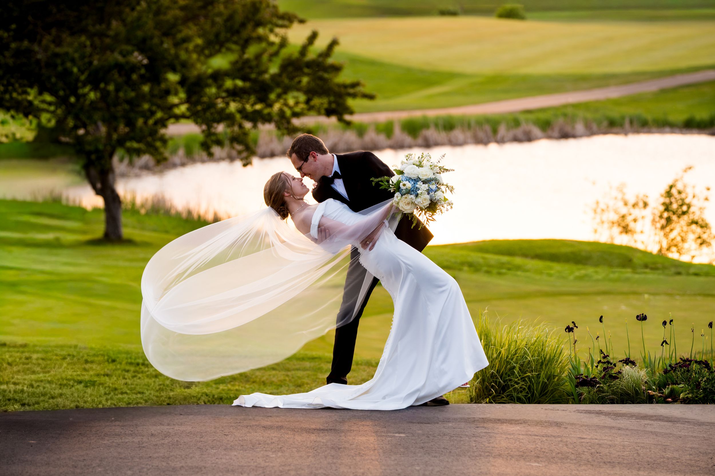 Bride and groom share a romantic dip kiss outdoors on a golf course with a scenic pond and green hills in the background.
