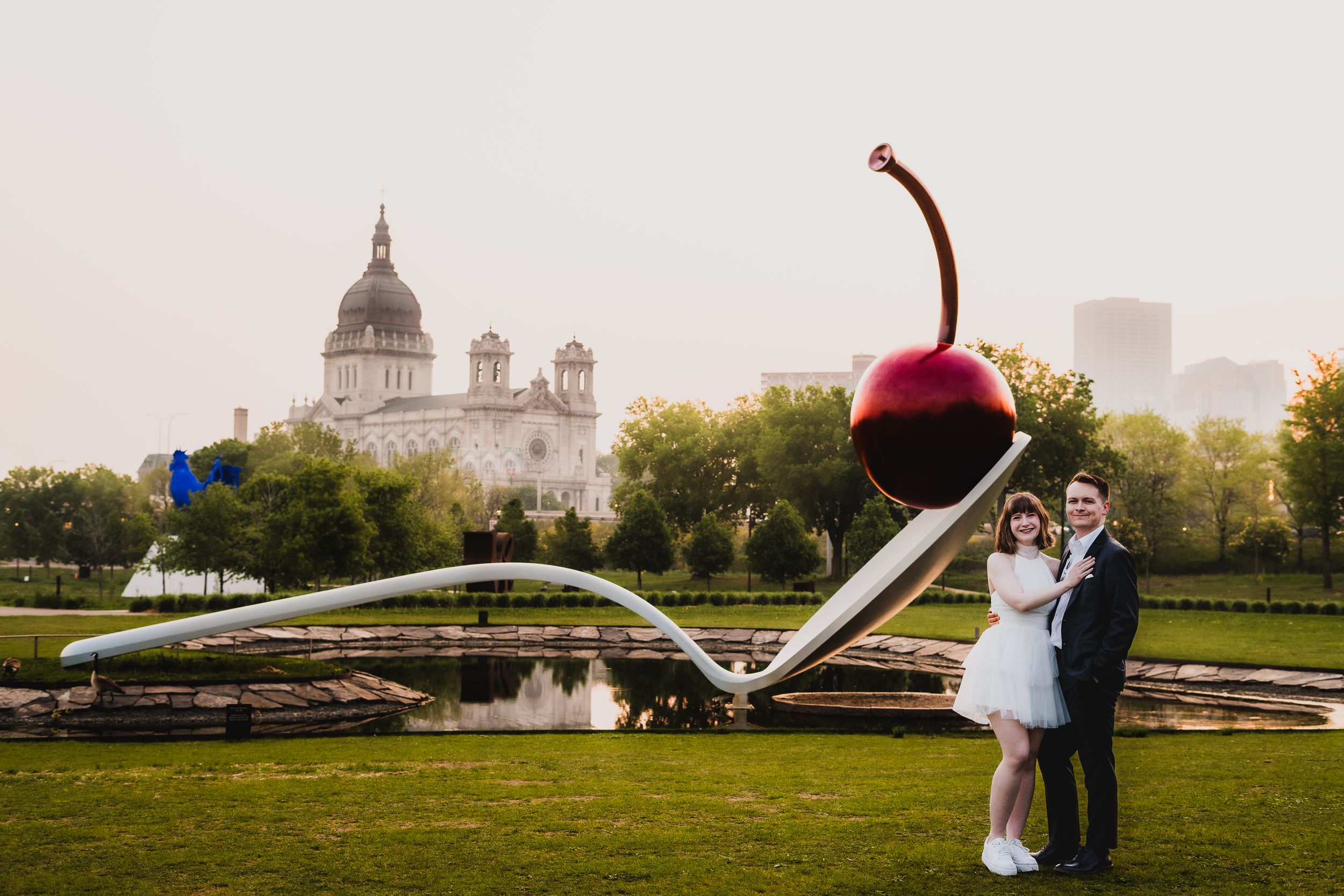 A couple poses near a giant cherry sculpture with the Minneapolis Sculpture Garden backdrop.