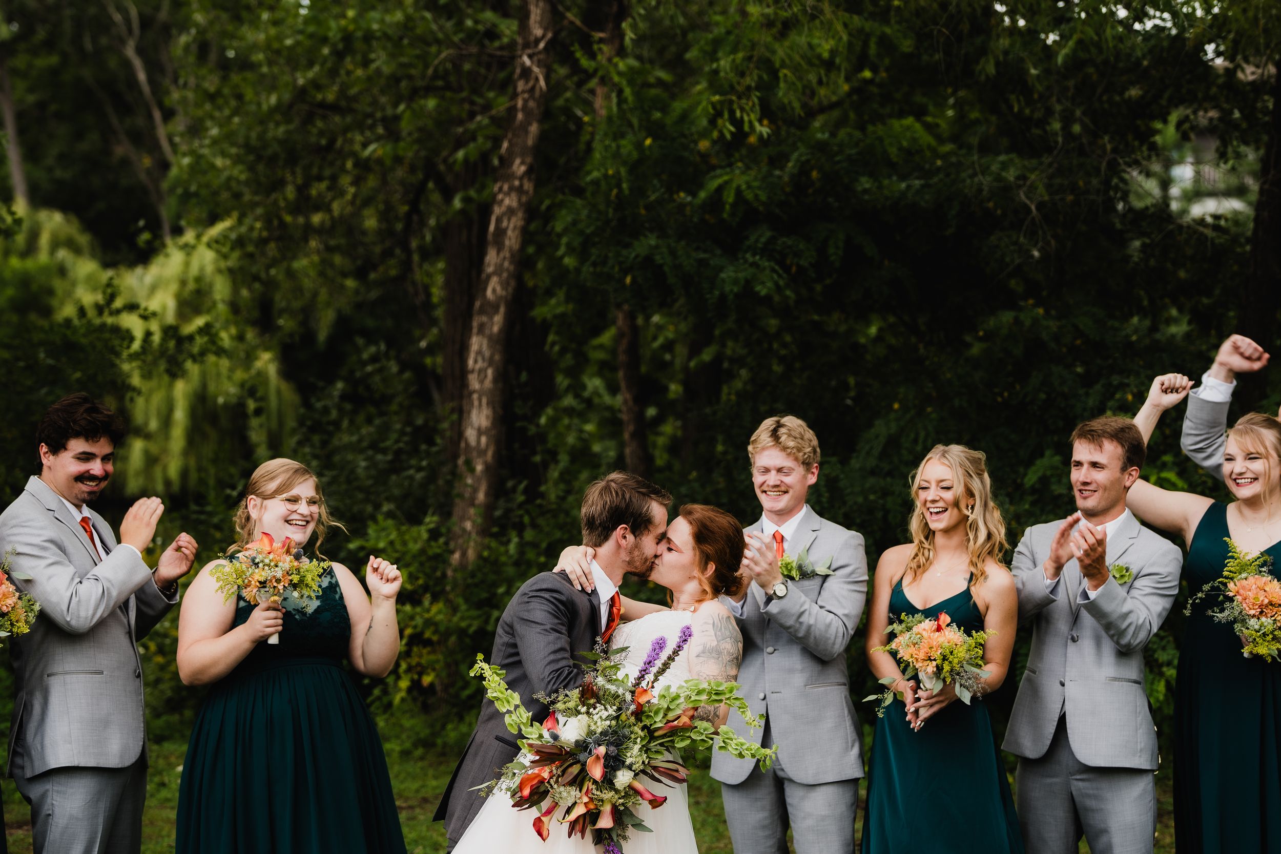 Wedding party in grey suits and dark green dresses pose for photos while holding colorful bouquets in an outdoor garden setting.