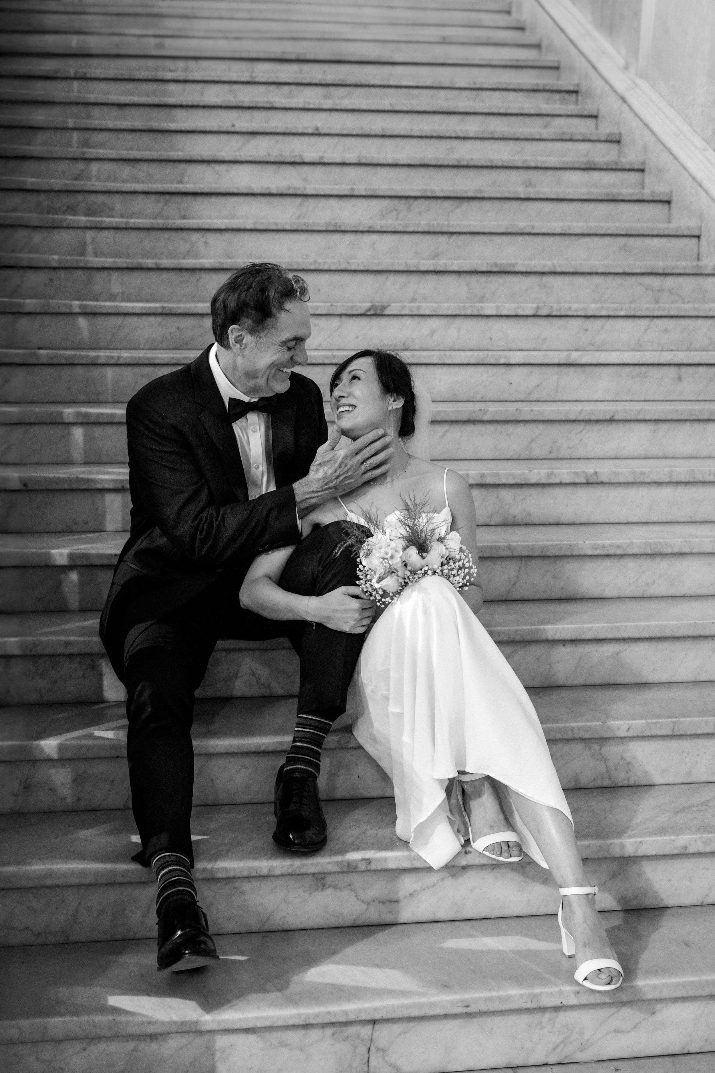 A black and white wedding photo of a couple sharing a joyful moment while sitting on elegant stone stairs.