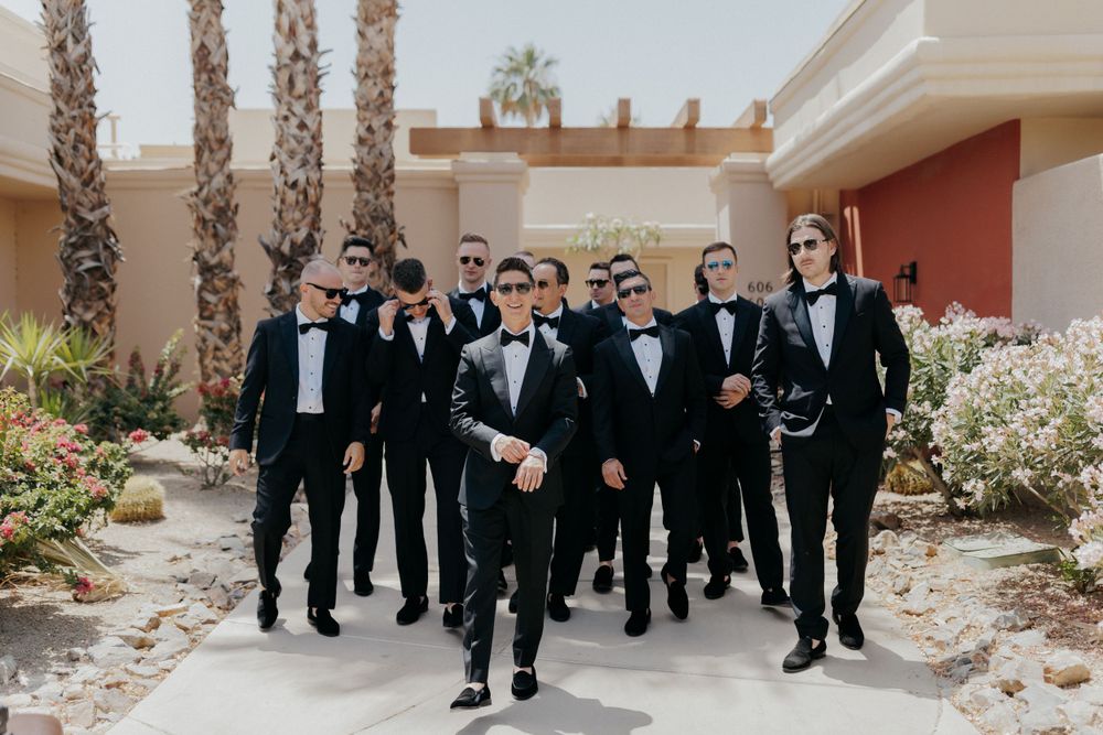 A group of groomsmen in black tuxedos walking together at a desert wedding venue.