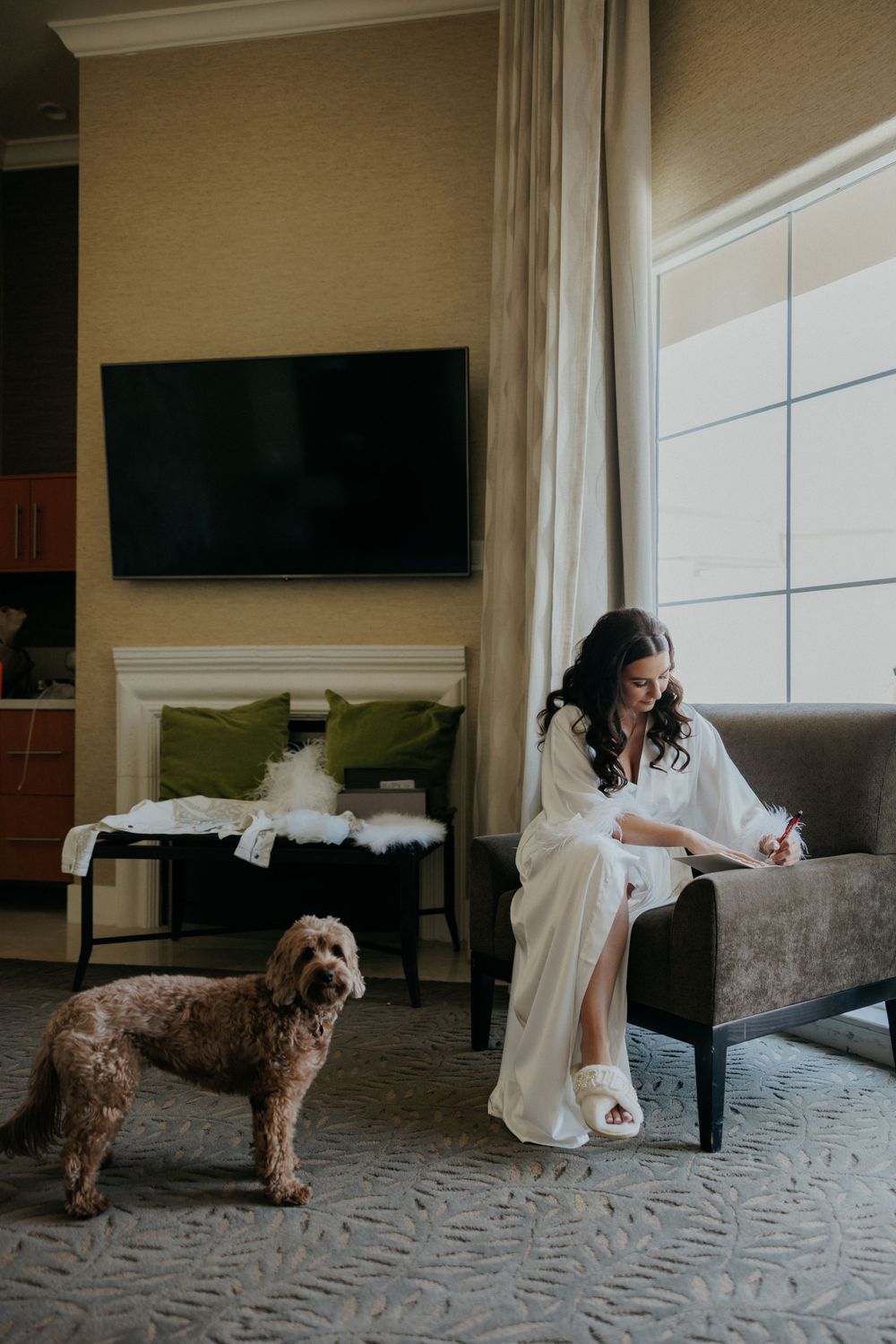A bride in a white robe sits on a gray couch with a brown goldendoodle dog in an elegant living room setting.