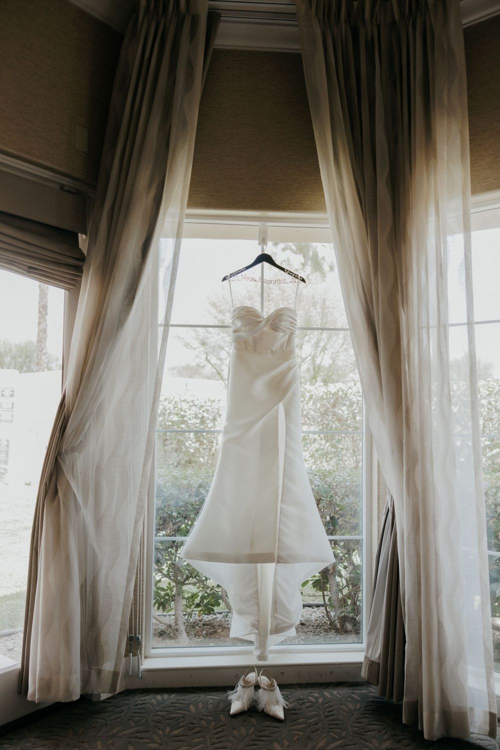 Wedding dress hanging in window frame with bridal shoes beneath in moody lighting.