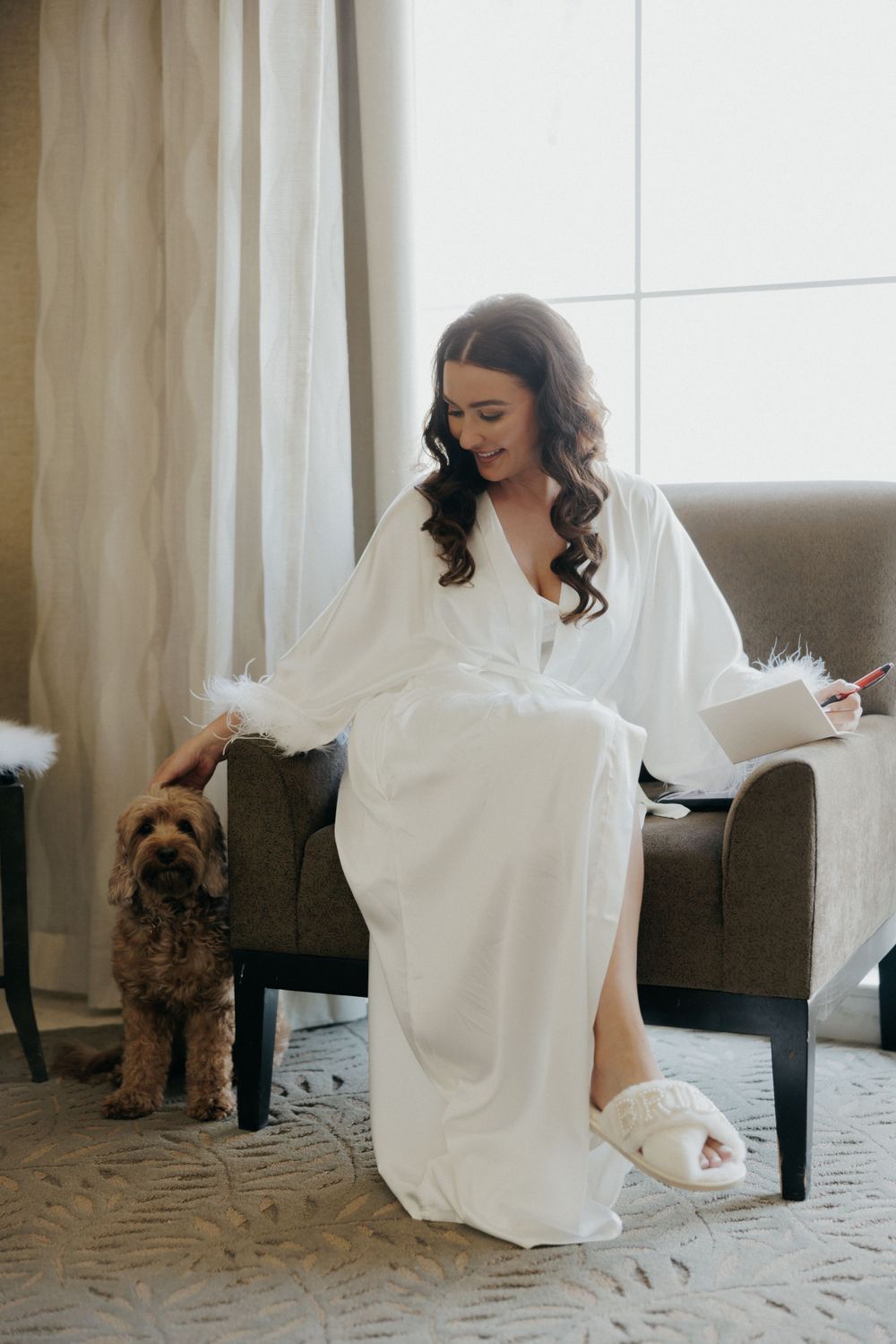 A person in a white robe sits in a gray armchair petting a small brown dog during wedding preparations.