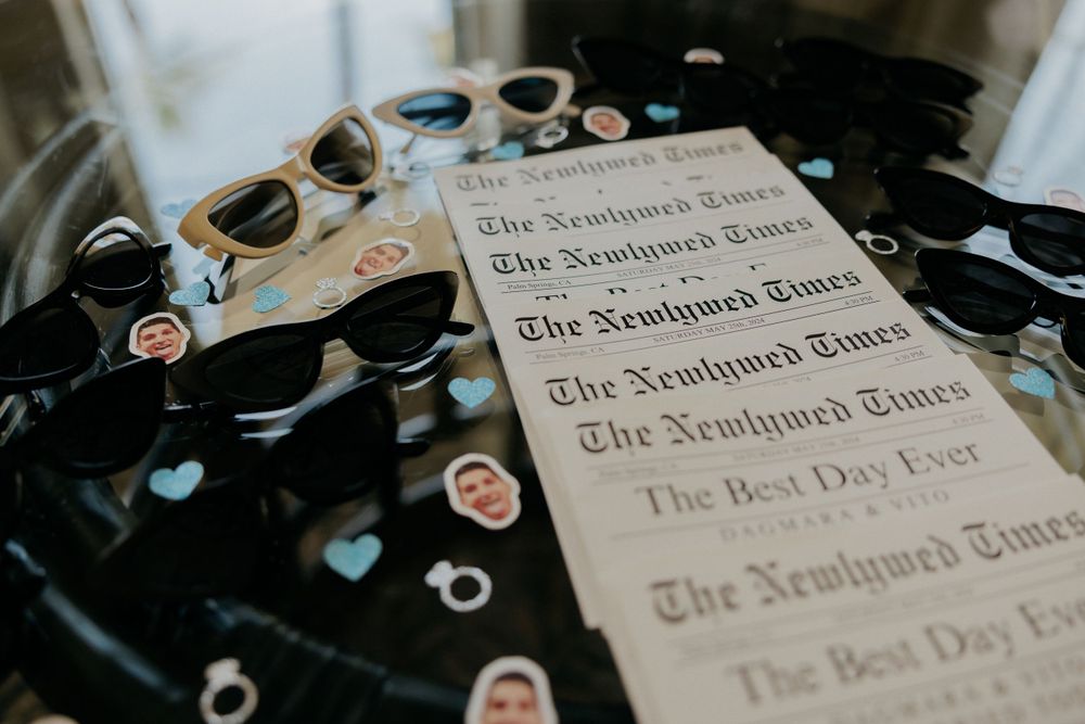 Wedding accessories including sunglasses and custom newspaper programs arranged on dark surface.