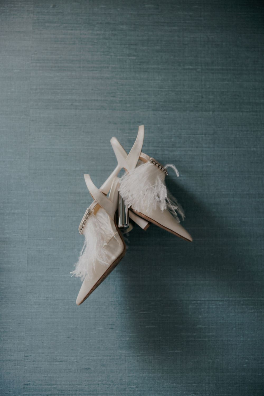 White feathered bridal shoes with delicate heels on a gray textured background.