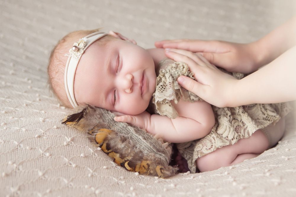 Peaceful newborn baby sleeping in lace outfit with floral headband, cradled gently by mother's hands on soft blanket.