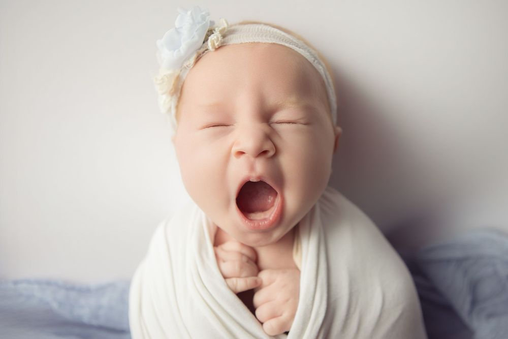Newborn baby girl yawning while swaddled in white wrap, wearing a delicate floral headband.
