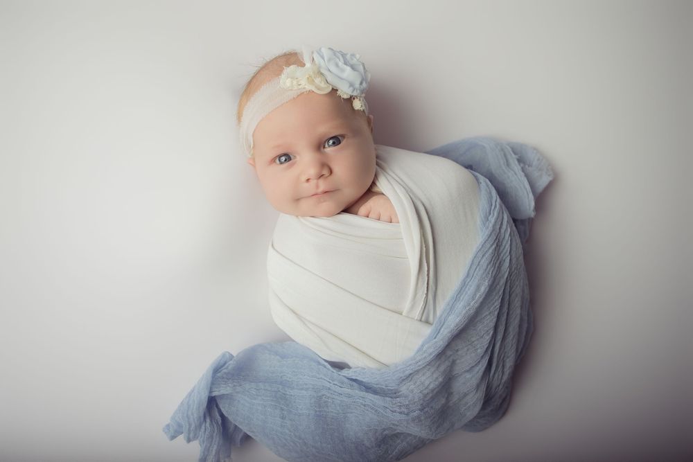 Newborn baby girl swaddled in white and blue wrap, wearing a floral headband, posed on a white background.