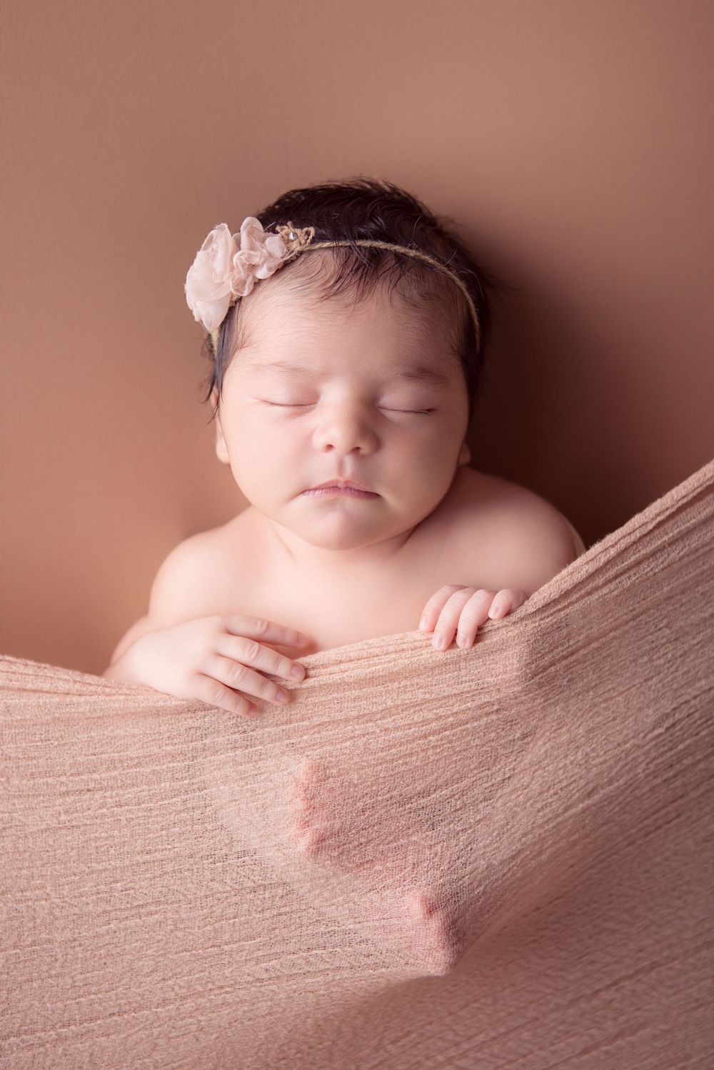 Sleeping newborn baby girl wearing a floral headband, wrapped in a soft pink cloth against a warm brown background.